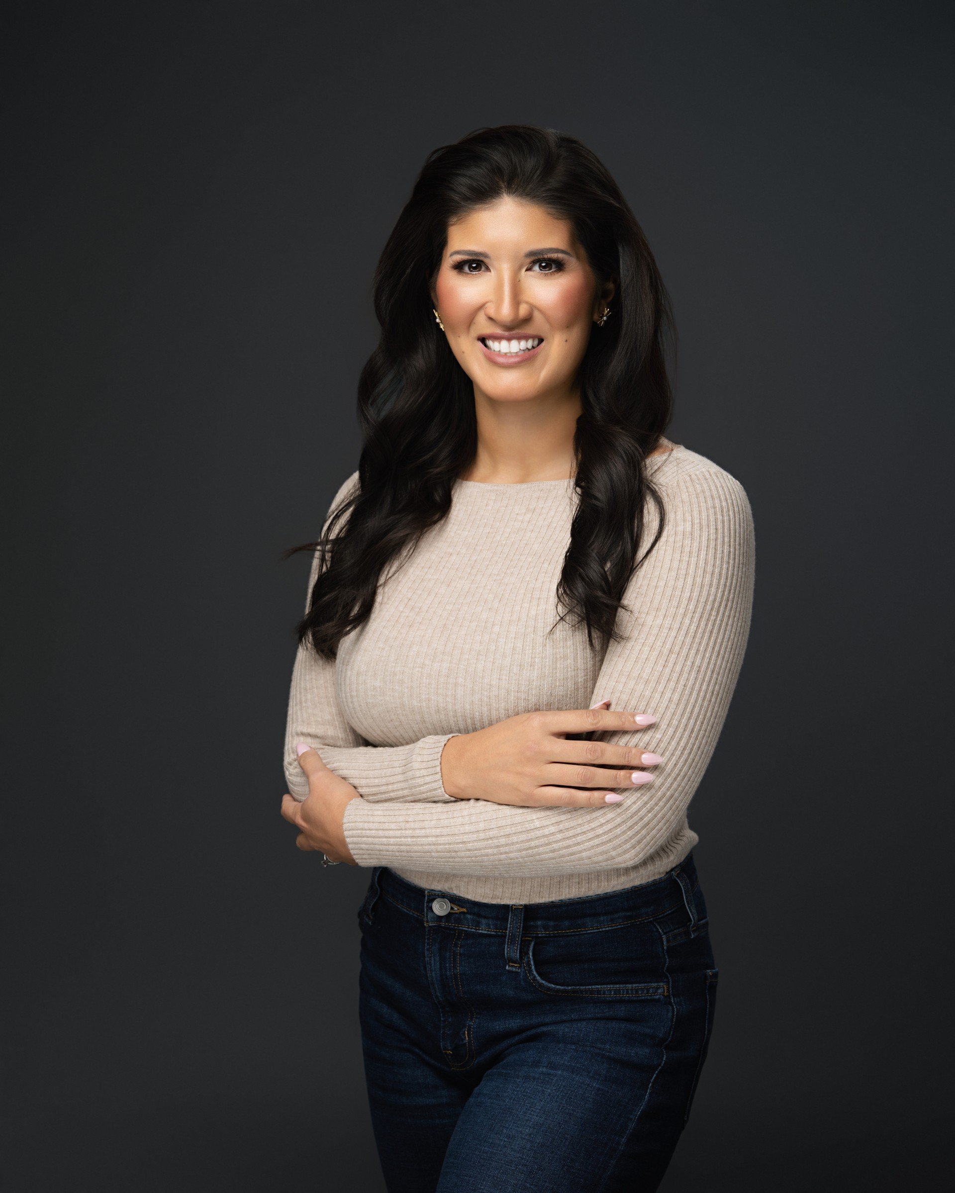 A woman with long, dark wavy hair smiling, wearing a beige ribbed sweater and dark jeans, standing against a dark grey background.