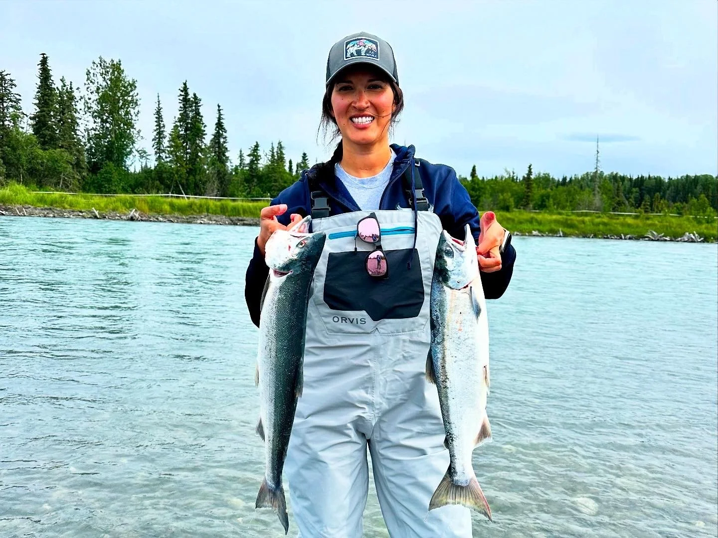 A smiling woman standing in a lake holding two large fish, one in each hand, with a forested shoreline in the background.