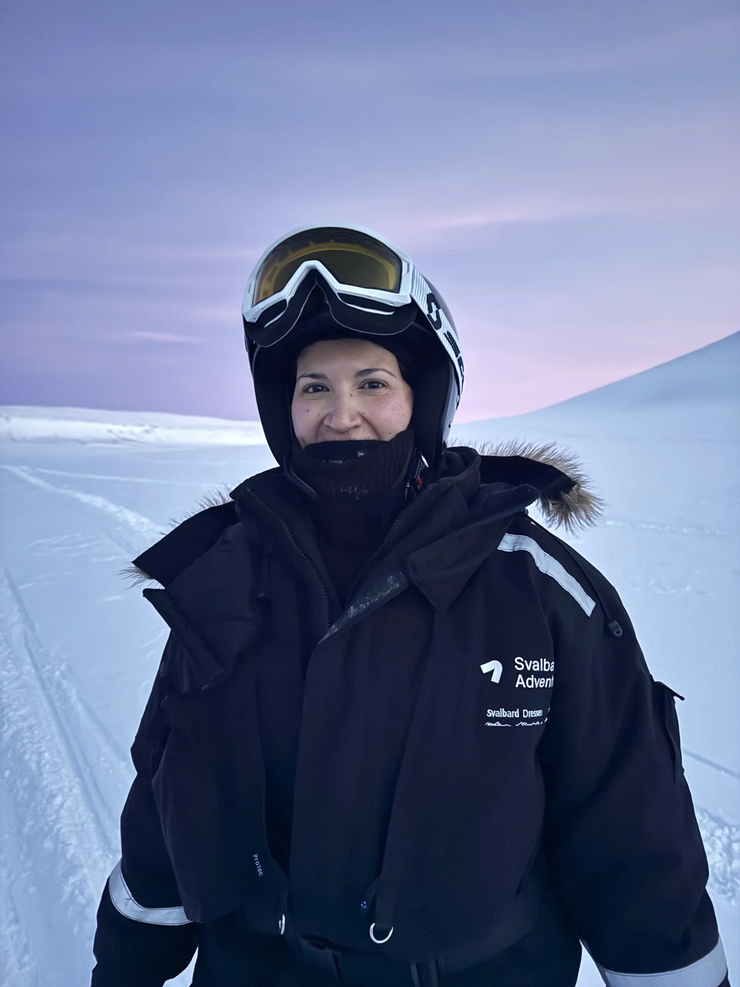 Woman in winter ski gear, including a black jacket with a logo, standing on snow with a mountain and pastel sky in the background.