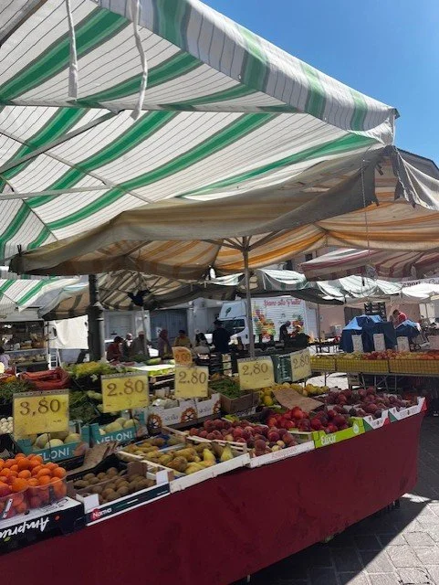 Outdoor fruit market with tables of fresh fruit under striped green and white canopies, with price signs and shoppers in the background.