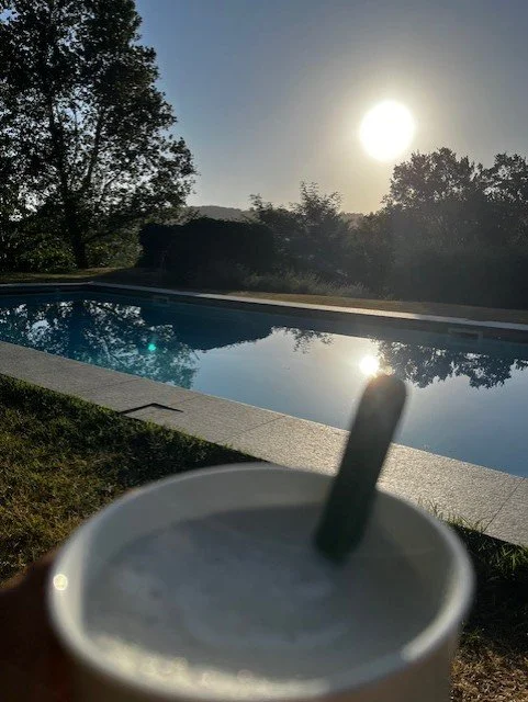 A swimming pool outdoors with the sun setting or rising in the background, and a blurry iced beverage in the foreground.