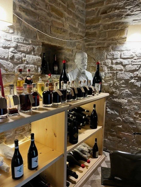 A small wooden shelf displaying various bottles of wine and liquor, with a stone wall background and a bust sculpture in the corner.