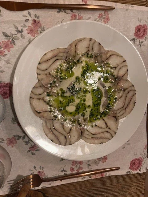A white plate with sliced, cooked octopus arranged in a circular pattern, garnished with green herbs and drizzled with olive oil.