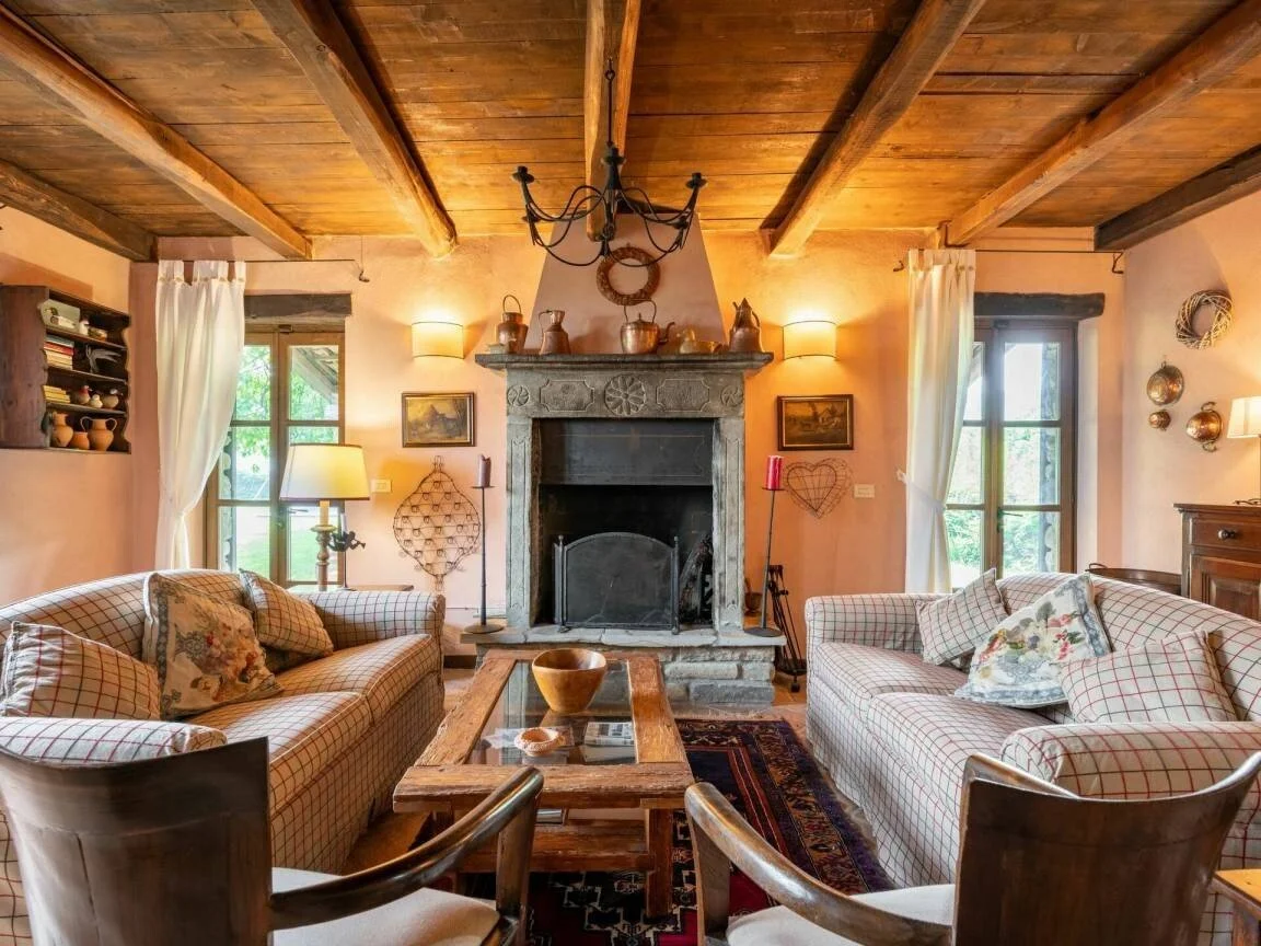 Living room with wood-beamed ceiling, stone fireplace, two plaid sofas with cushions, wooden coffee table, side tables with lamps, windows with curtains, and various decorative items.
