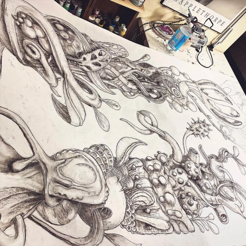A detailed black and white sketch of various abstract and biological shapes on a large sheet of paper, with art supplies and a water bottle in the background.