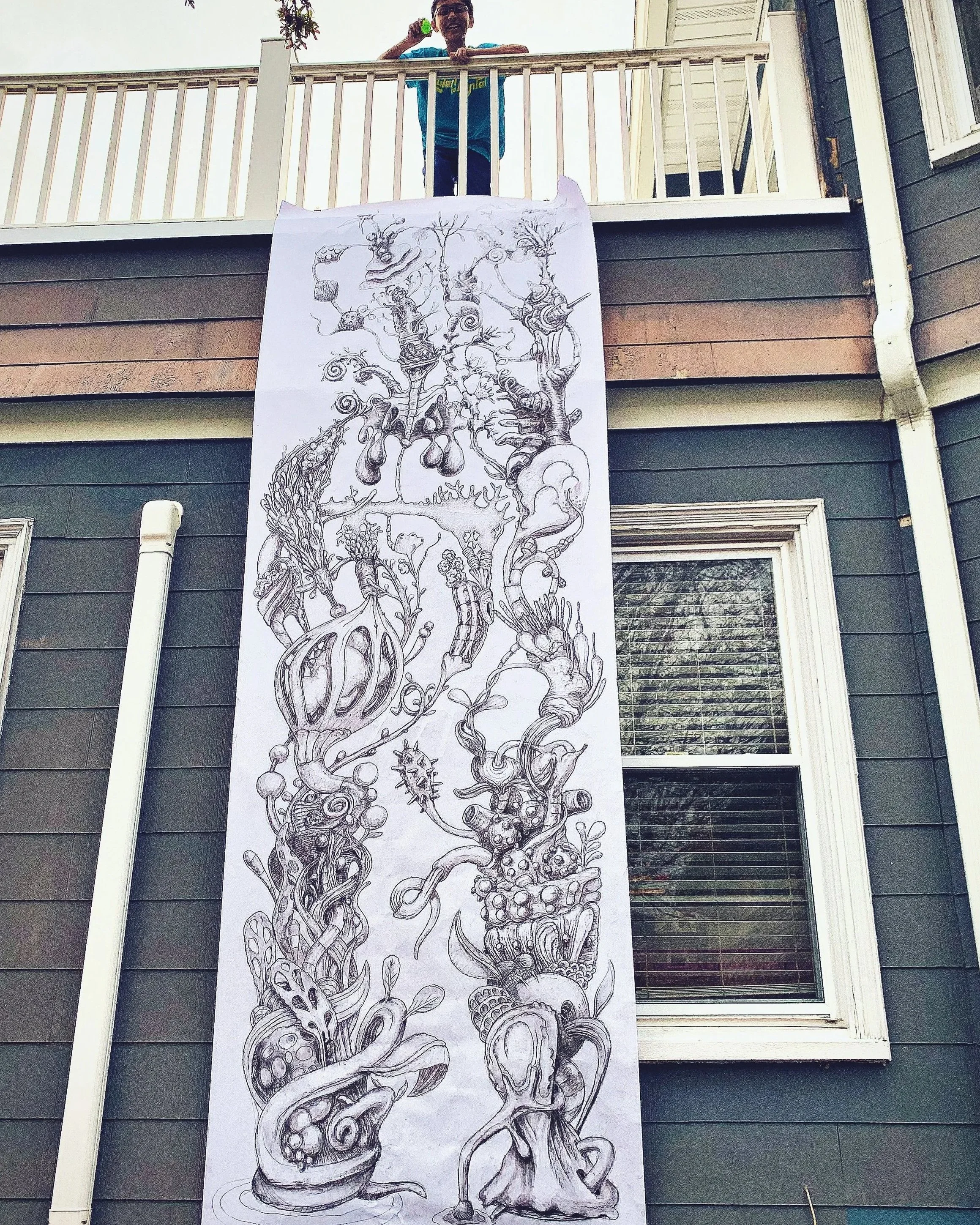 A large, detailed black and white drawing hanging on the side of a house, with a person standing on a balcony above it. The drawing features a surreal, intricate composition of various abstract and organic shapes, including faces, plants, and fantast