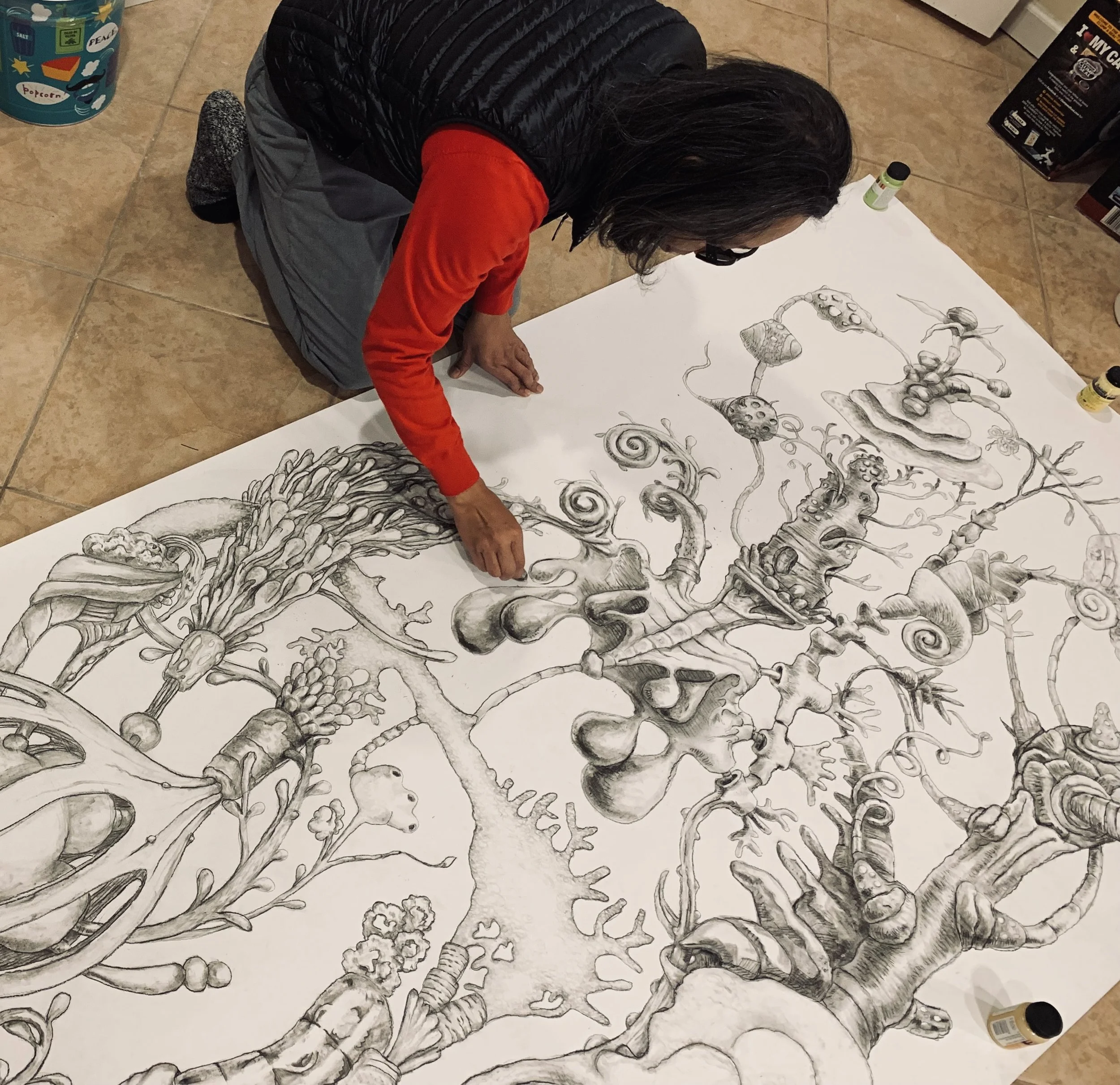 Person working on a large detailed pencil drawing of a fantastical tree with various whimsical and organic shapes on a white sheet on the floor.