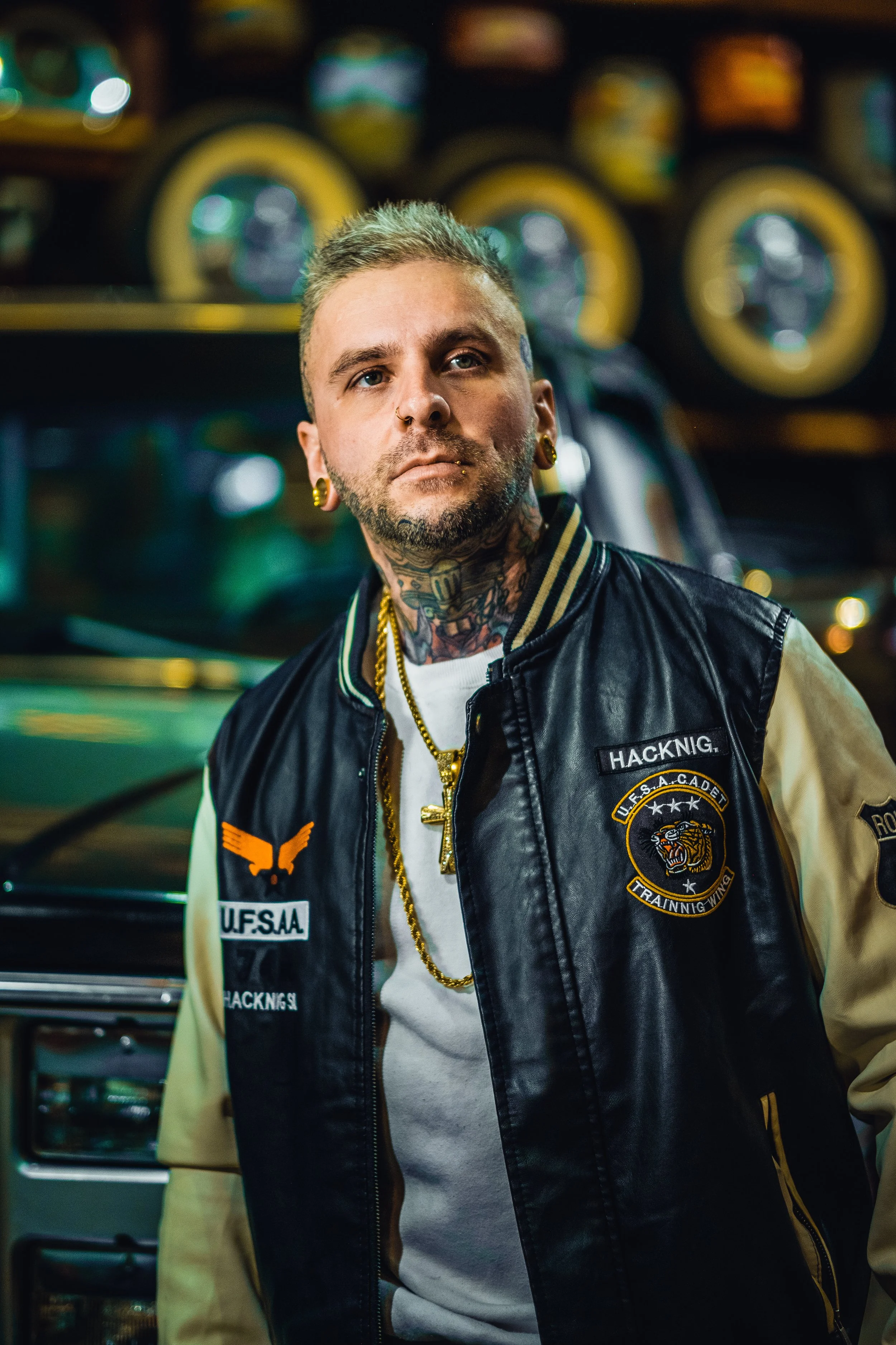 A young man with tattoos and piercings wearing a black and beige varsity jacket with patches, standing in front of a vehicle at night.