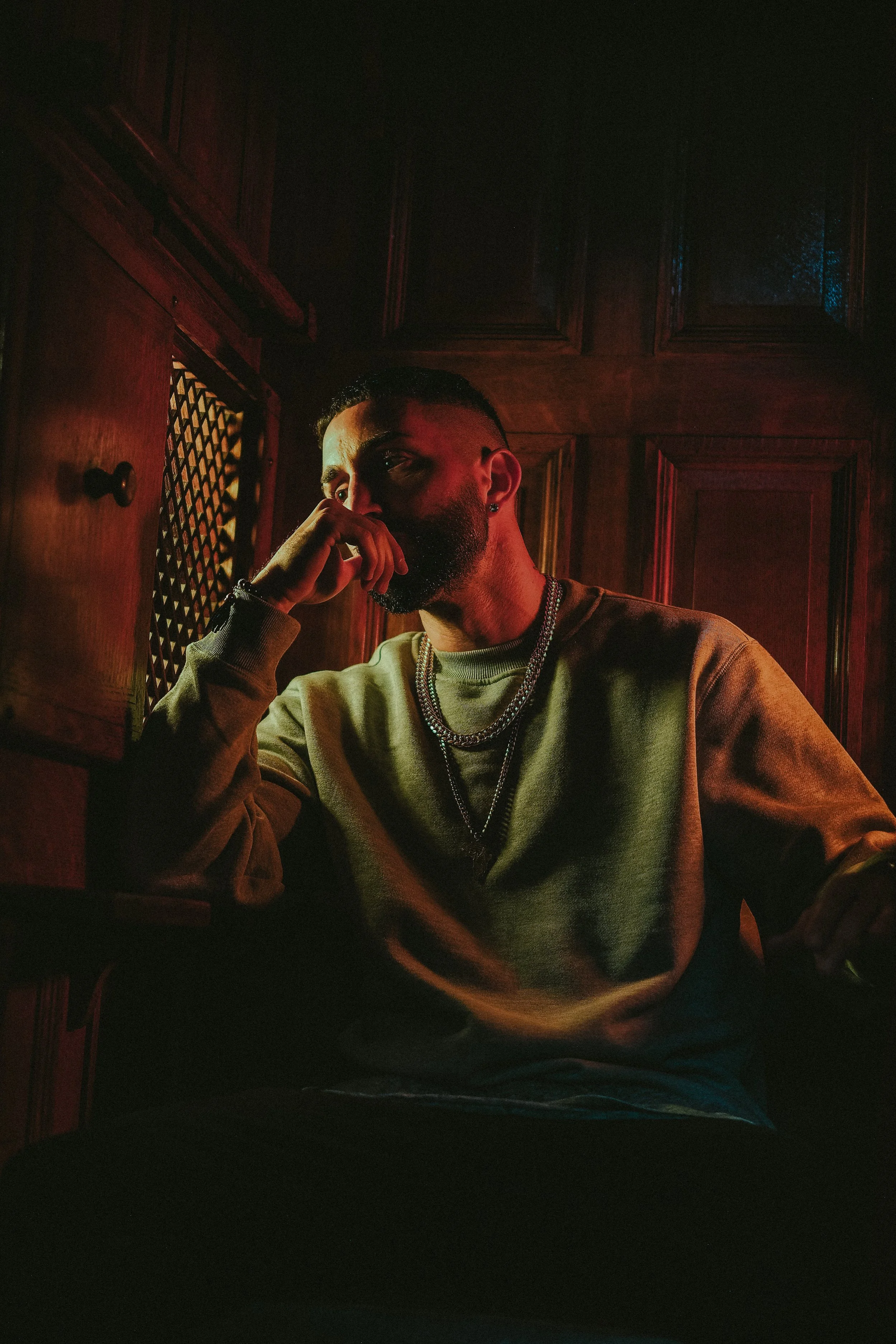 Videographer in Montreal capturing professional music videos, corporate videos, and livA man with a beard and short hair sitting in a dimly lit room with wooden walls, wearing a beige sweatshirt and layered chains, looking thoughtfully at the camera.