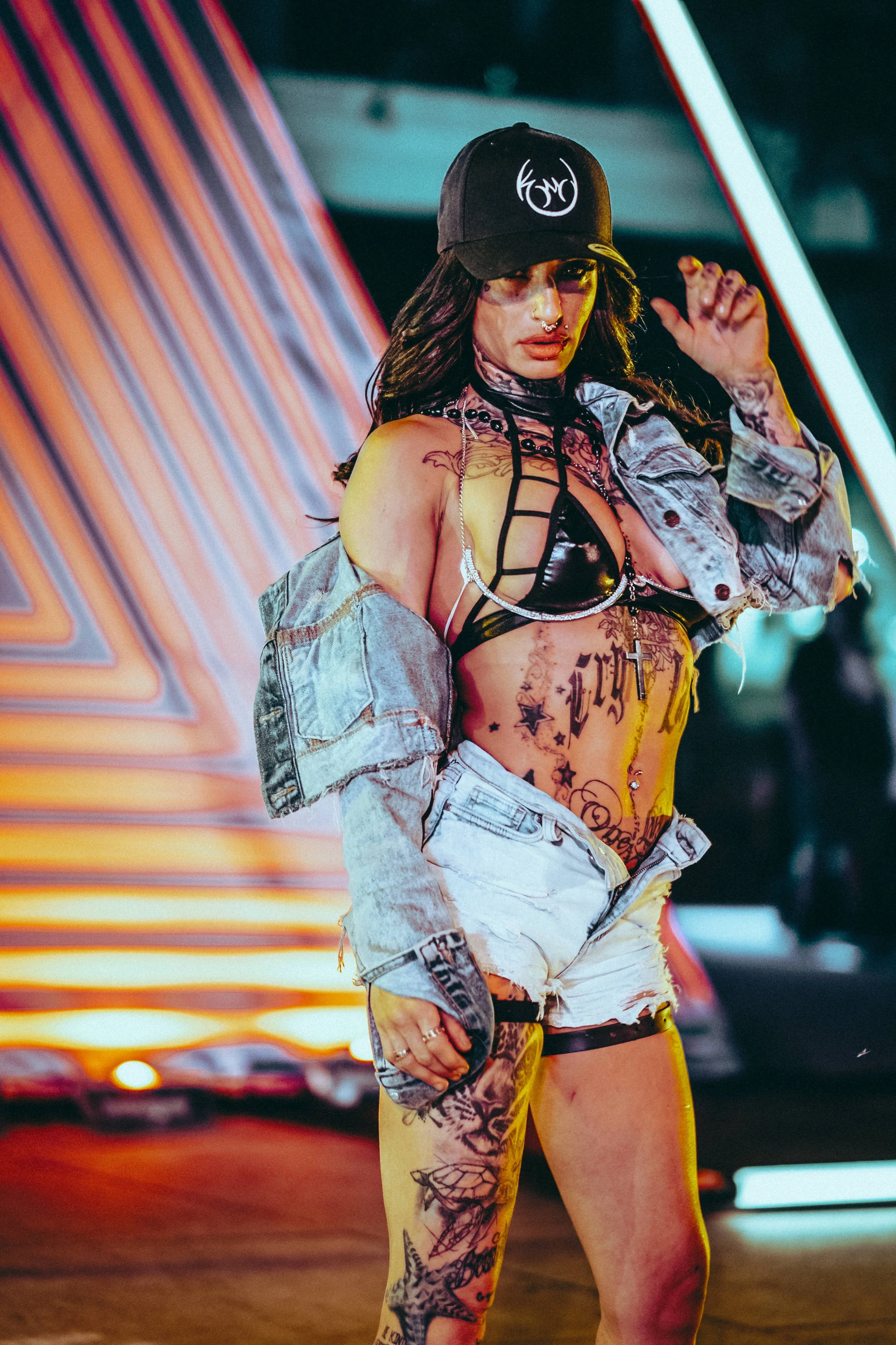 Videographer in Montreal capturing professional music videos, corporate videos, and live events. High-quaA woman in a baseball cap, denim jacket, and lingerie top with tattoos, posing with her hand near her head against colorful, neon-lit background.