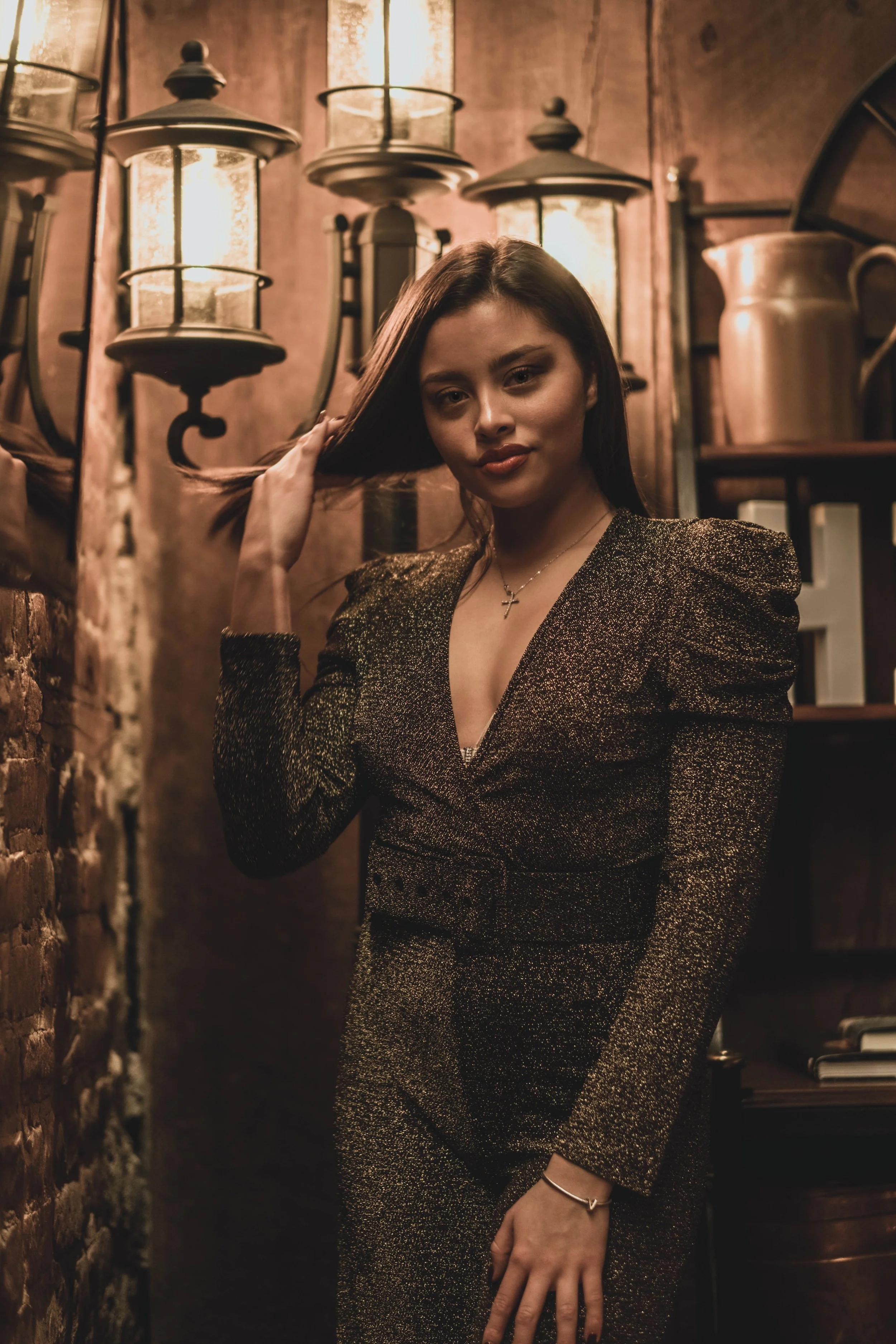 A woman in a sparkly black and gold suit standing in a warmly lit, rustic environment with brick wall and vintage lanterns, touching her hair with one hand.