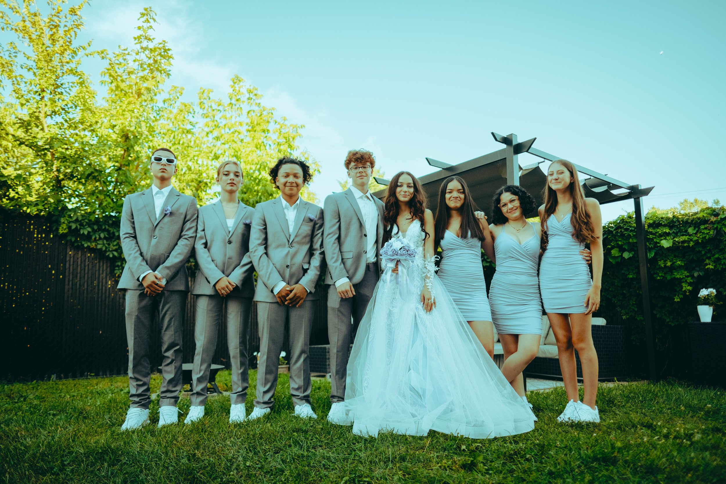 Videographer in Montreal capturinGroup of young adults dressed in formal attire, including a bride in a white wedding gown holding a bouquet, standing outdoors on grass with green trees and a modern outdoor structure in the background during daytime.