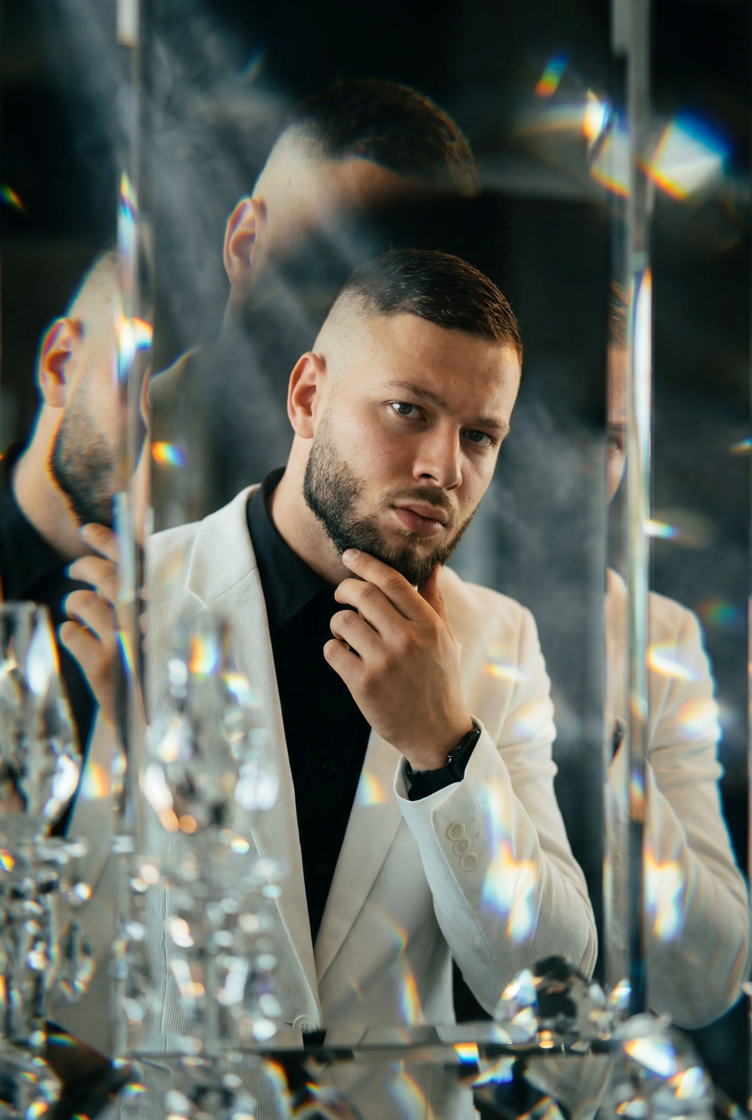 A young man with short dark hair, beard, and light skin looks thoughtfully into a mirror, resting his chin on his thumb and index finger, wearing a cream blazer over a black shirt, surrounded by crystal decorations and reflections in a dimly lit room