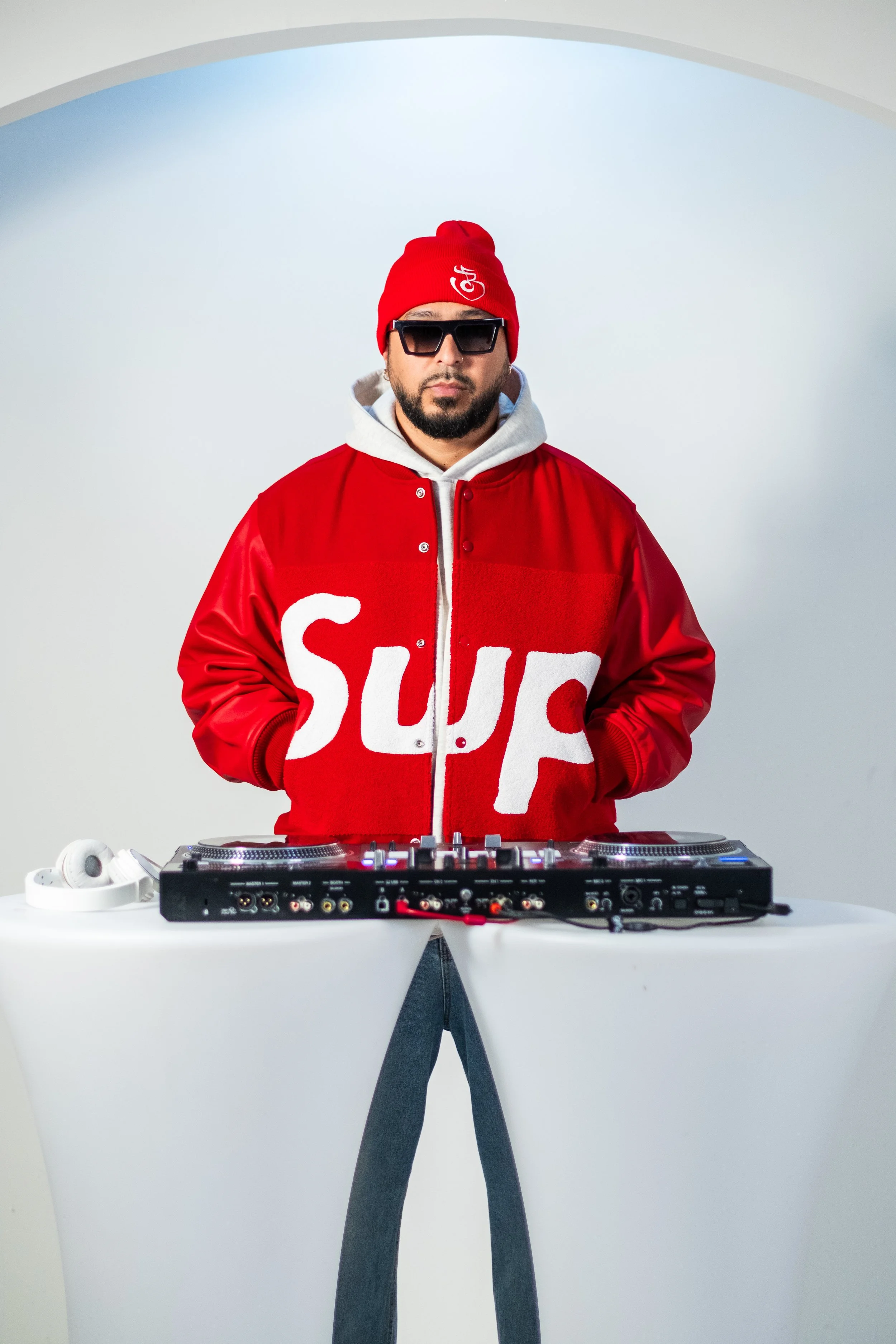 A man wearing a red beanie, sunglasses, and a red varsity jacket with white letters is standing behind DJ equipment with a white background.