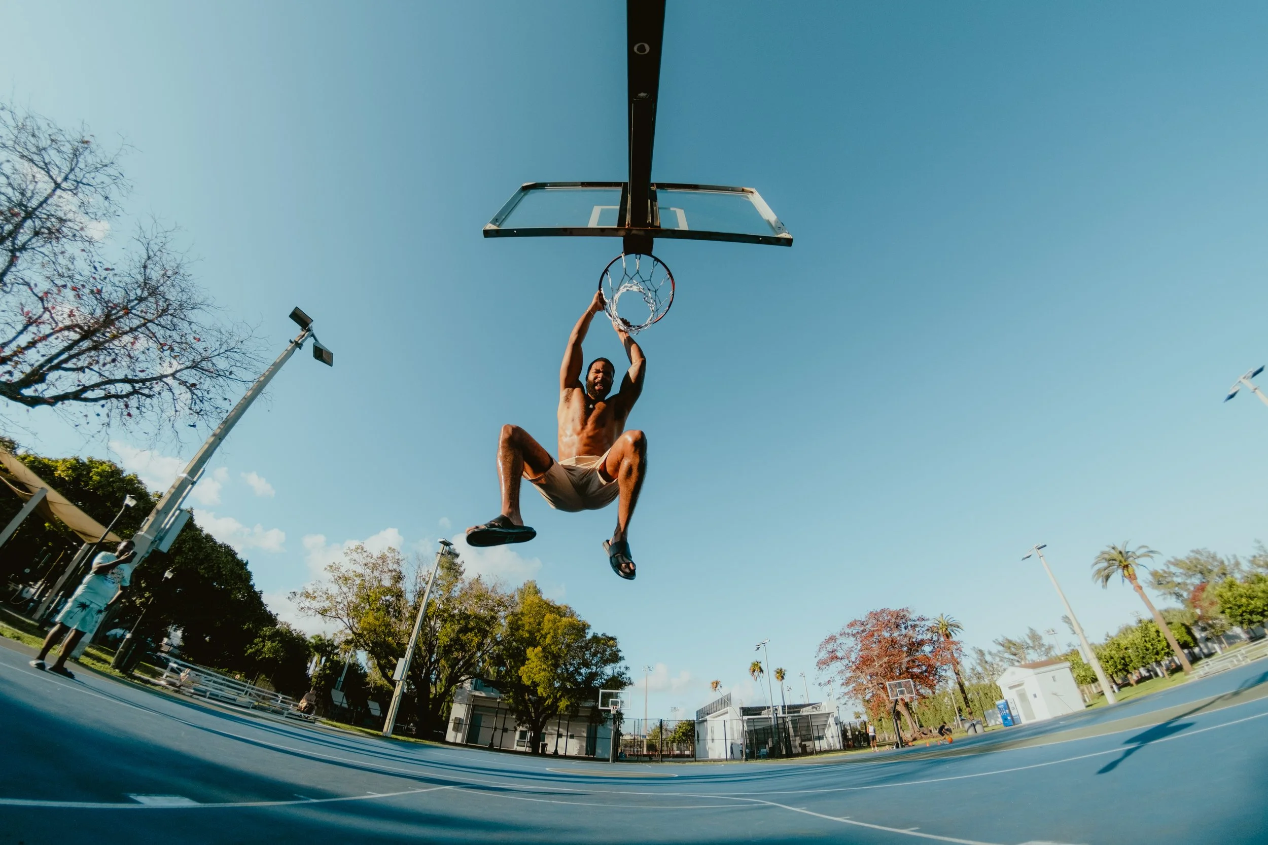 A man hanging from a basketball hoop on an outdoor court, mid-air, during daytime.