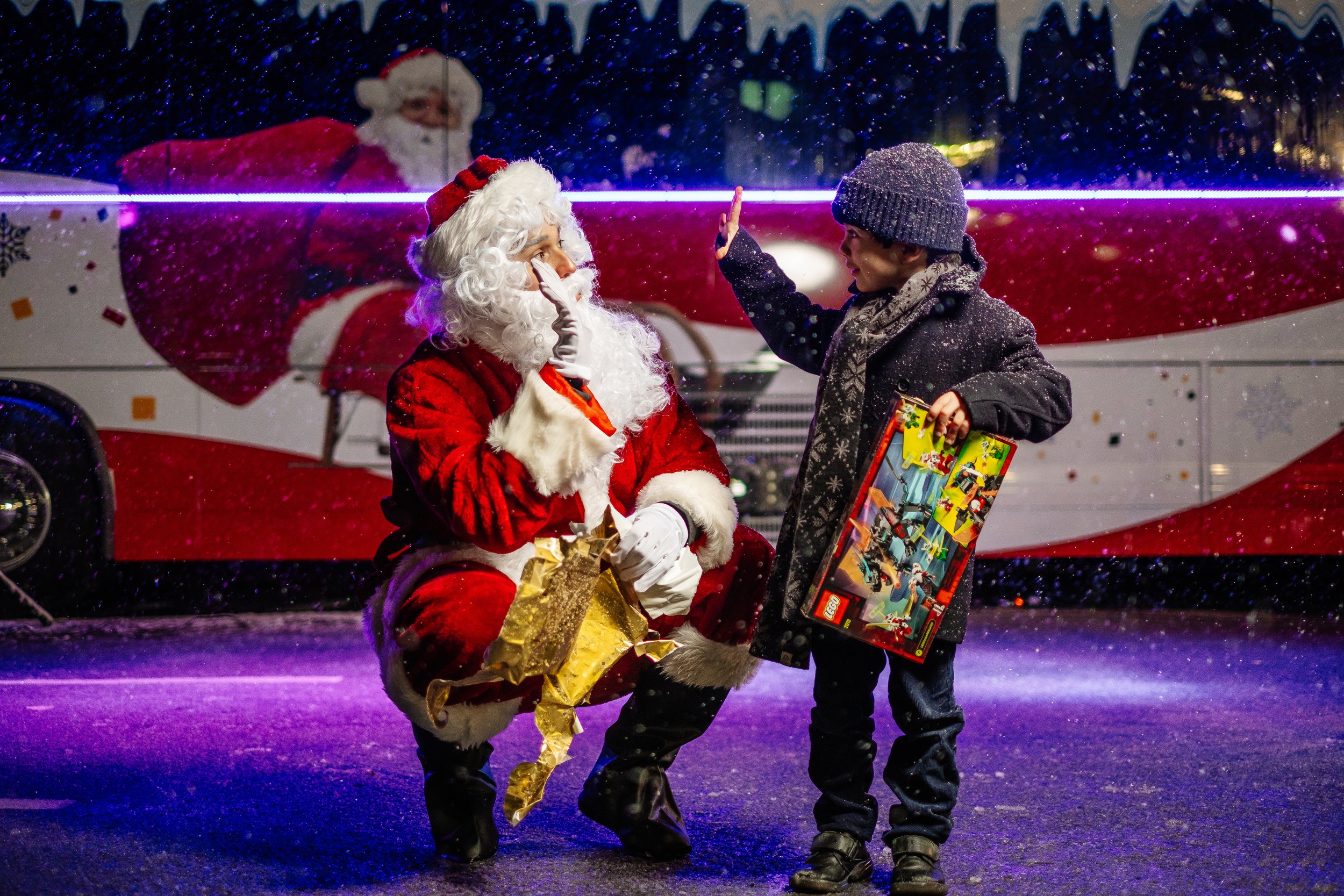Videographer in Montreal capturing professional music viA young boy giving a high five to Santa Claus during a festive outdoor scene at night with snow falling. Santa is seated with a gold gift bag, and there is a decorated vehicle in the background.