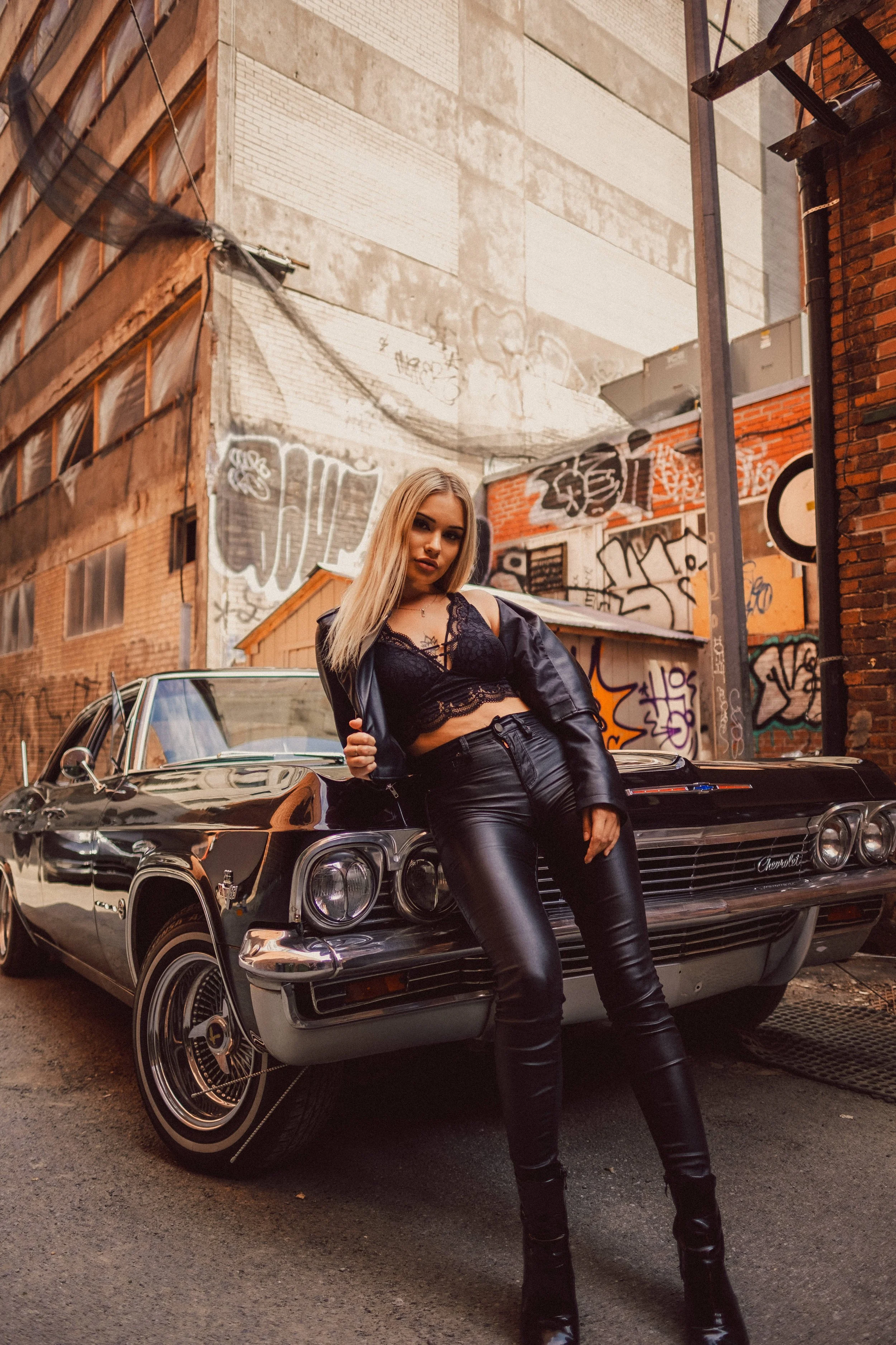 A woman with blonde hair standing next to a vintage black Chevrolet car in an alley with graffiti-covered brick walls.