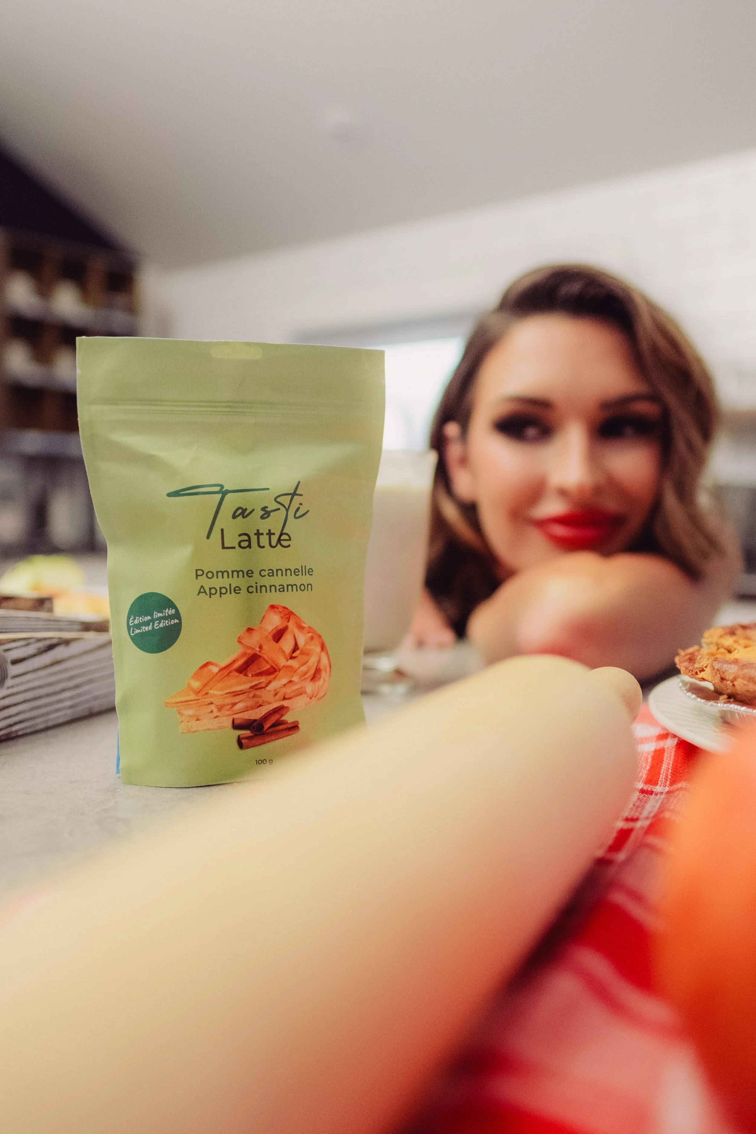 Videographer in Montreal capturing proClose-up of a green package of Tausti Latte with apple cinnamon flavor, with a woman with brown hair and red lipstick in the background, smiling and resting her chin on her arm, with a kitchen setting behind her.