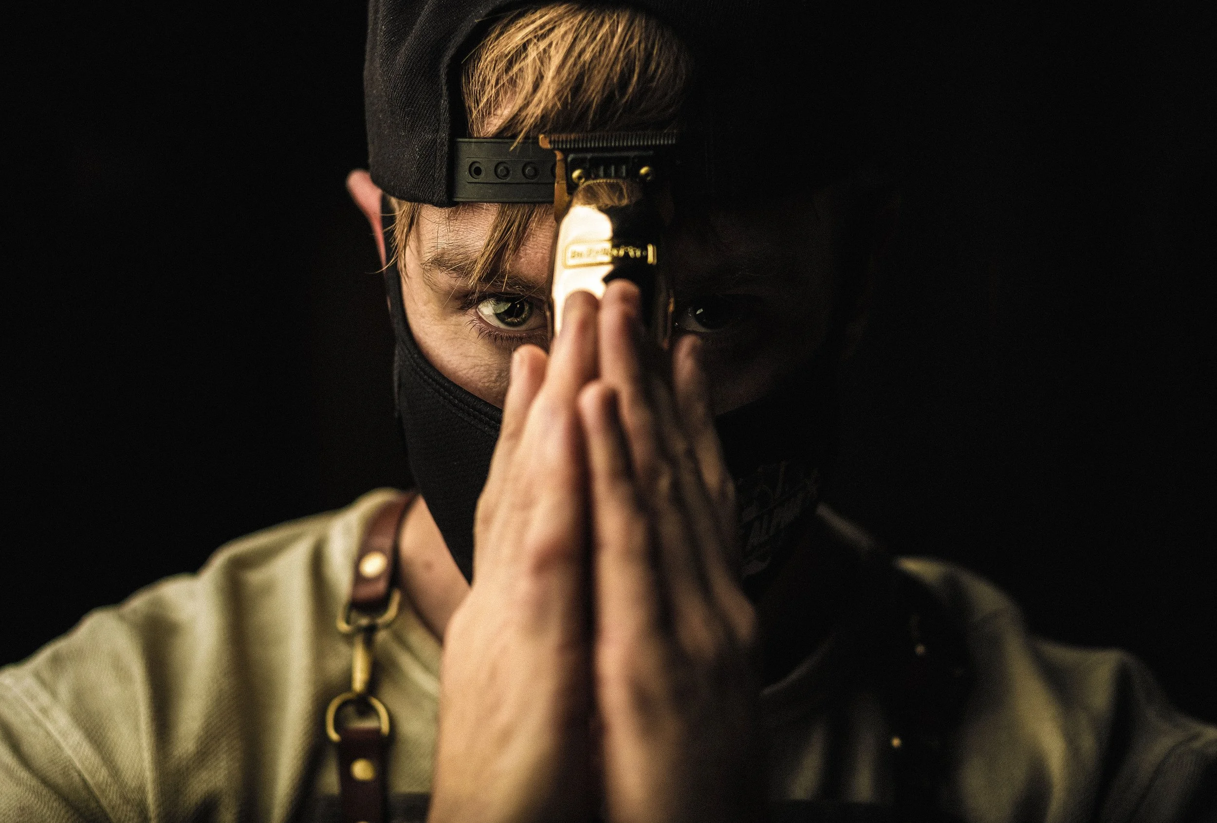 Person wearing a black cap and mask, holding a gun pointed directly at the camera, with intense green eyes visible.