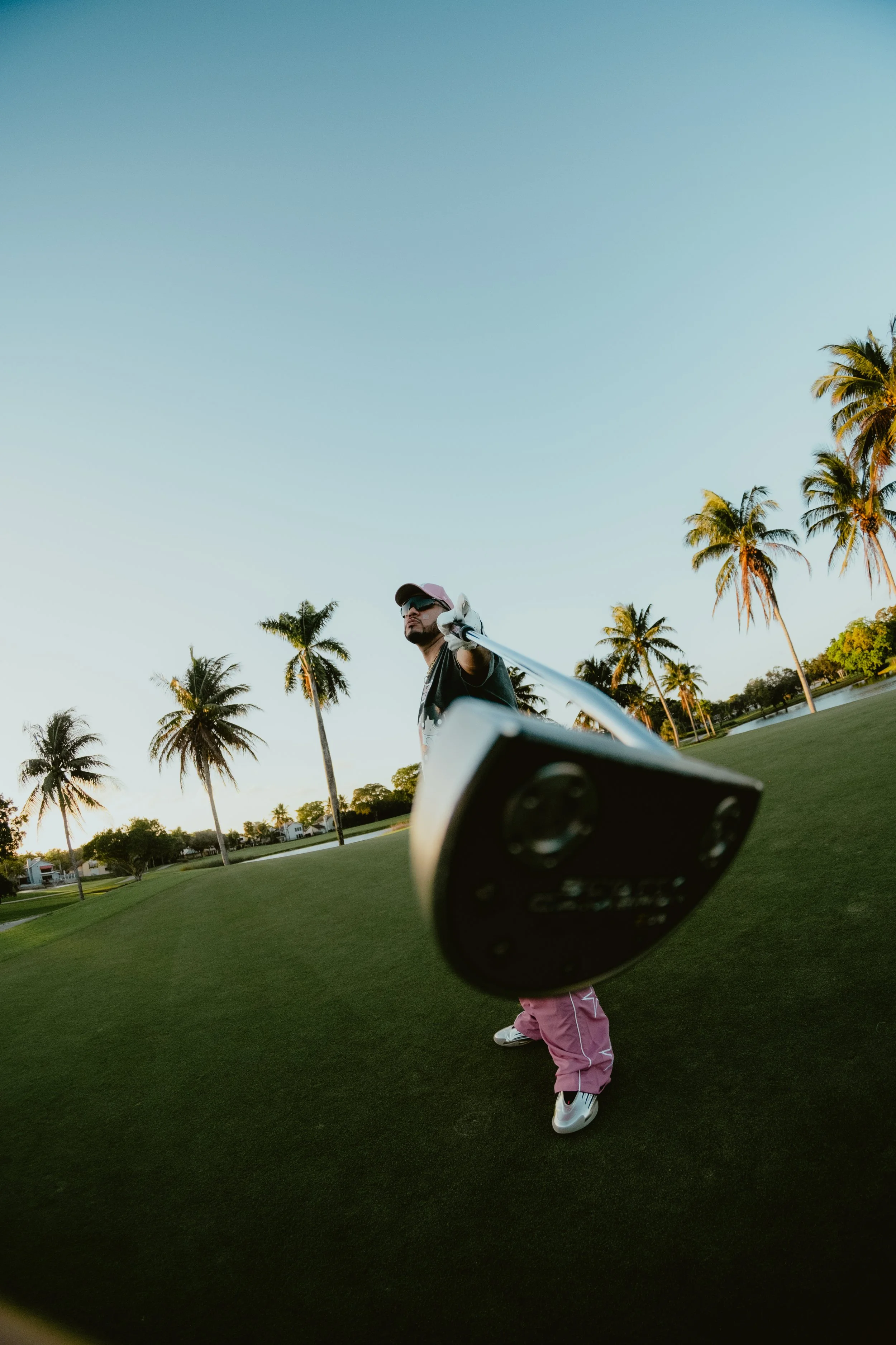 A man on a golf course swings a golf club, with the photo taken from a low angle near the ground, showing palm trees and a clear sky in the background.