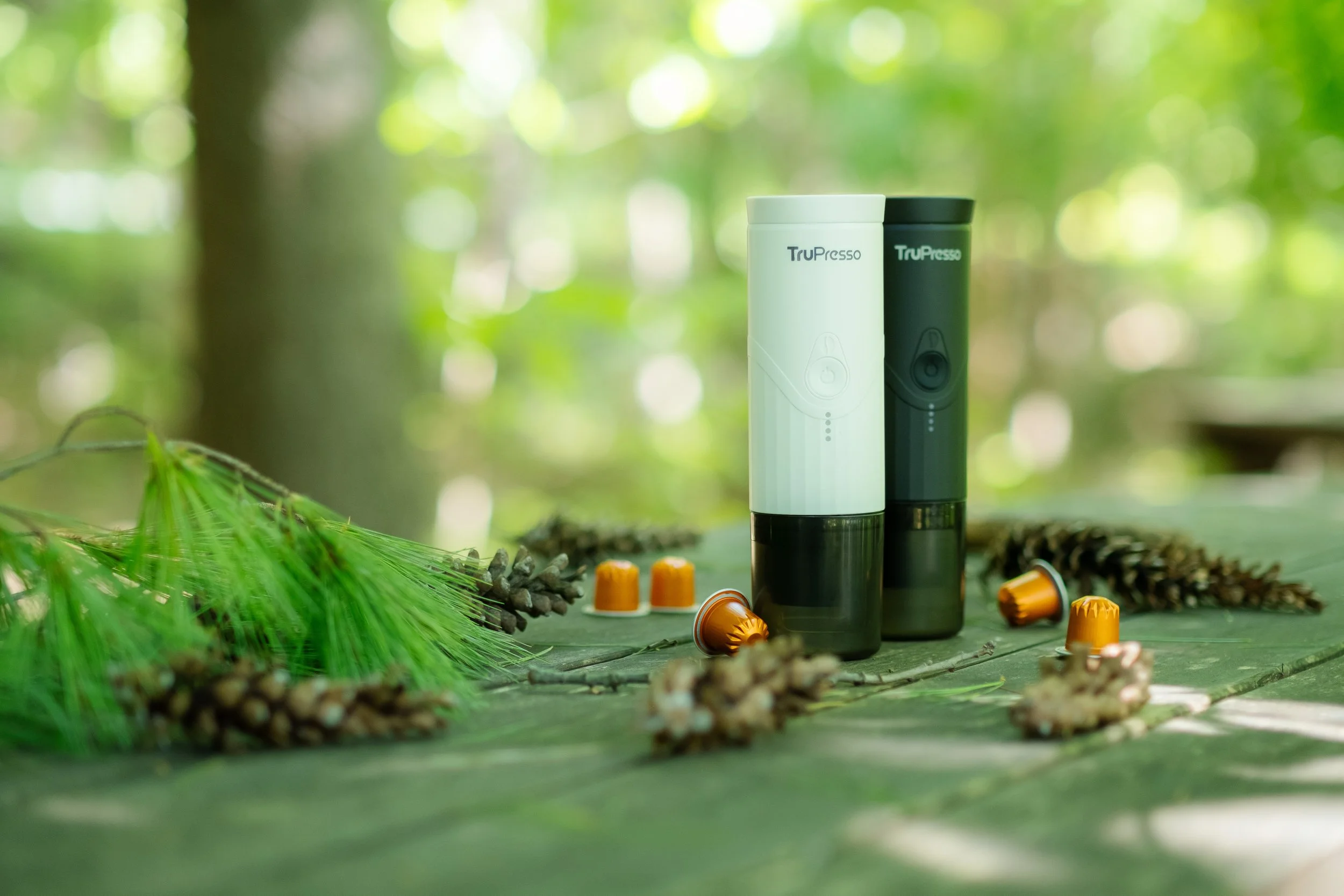 Videographer in Montreal capturing professional musTwo portable coffee makers, one white and one black, stand upright on a wooden surface outdoors. Surrounding them are coffee capsules and pine cones, with green foliage and blurred forest background.