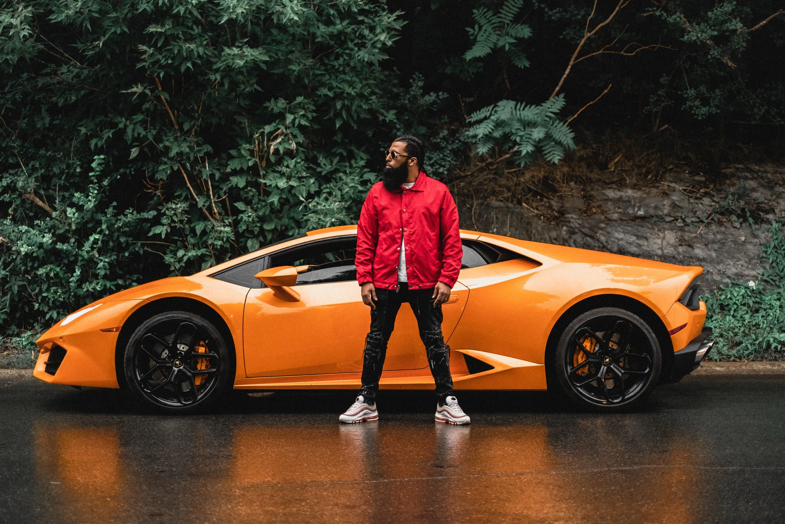 Videographer in Montreal capturing professional music videos, corporate videos, and live A man with a beard wearing sunglasses, a red jacket, and black pants stands in front of an orange sports car on a wet road, with green foliage in the background.