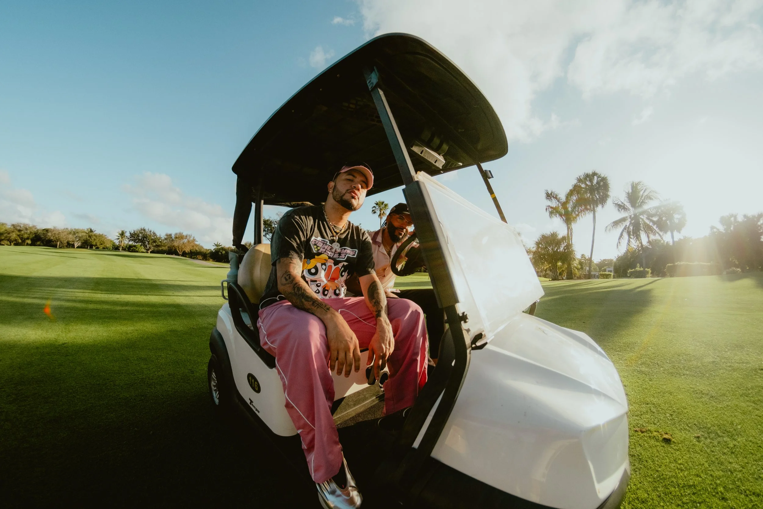 Two men sitting in a white golf cart on a golf course with palm trees in the background during sunset.