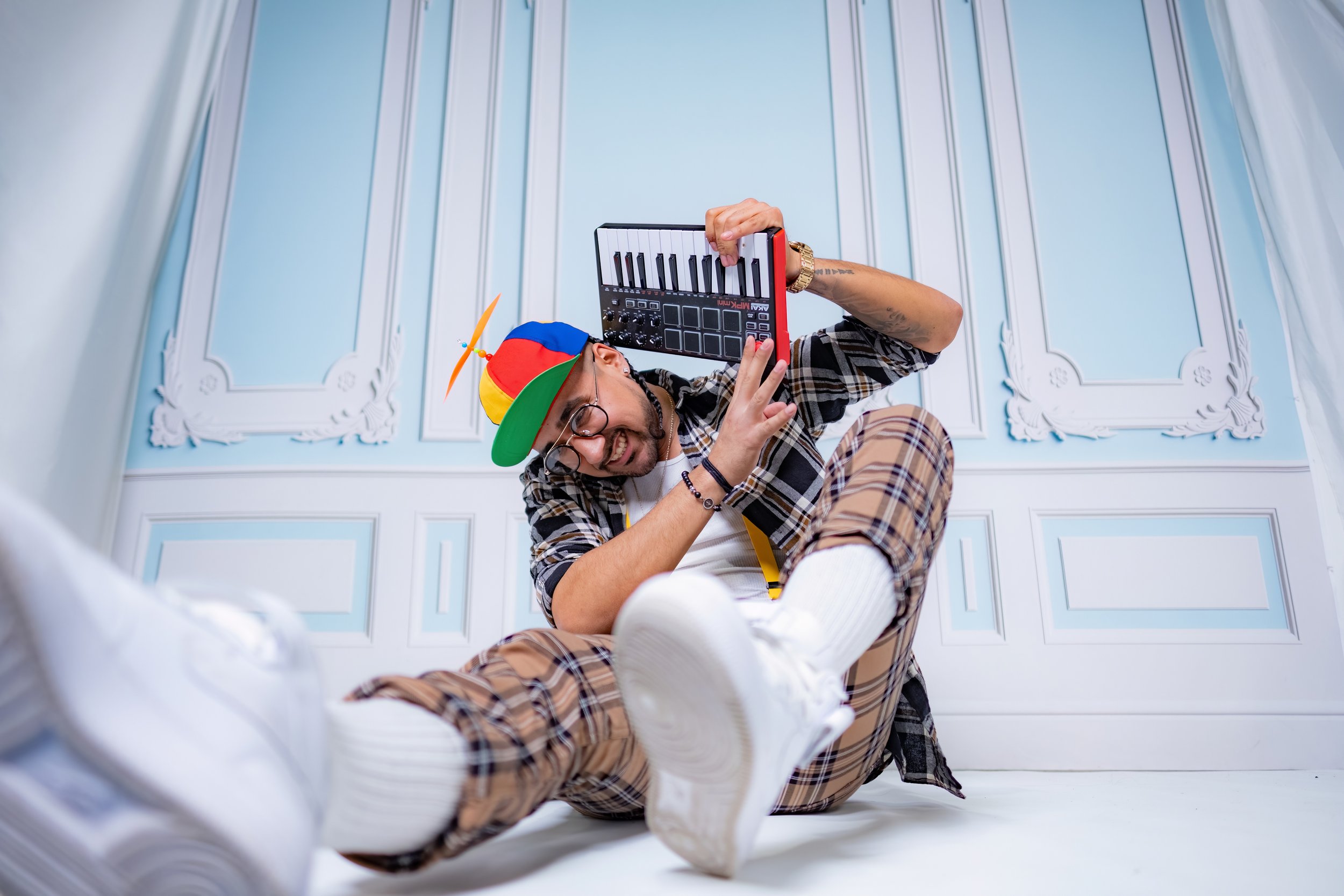 A young man wearing a colorful cap, glasses, plaid shirt, and checkered pants sitting on the floor in front of a light blue and white decorative wall, holding a music production controller, smiling, with a playful and energetic pose.