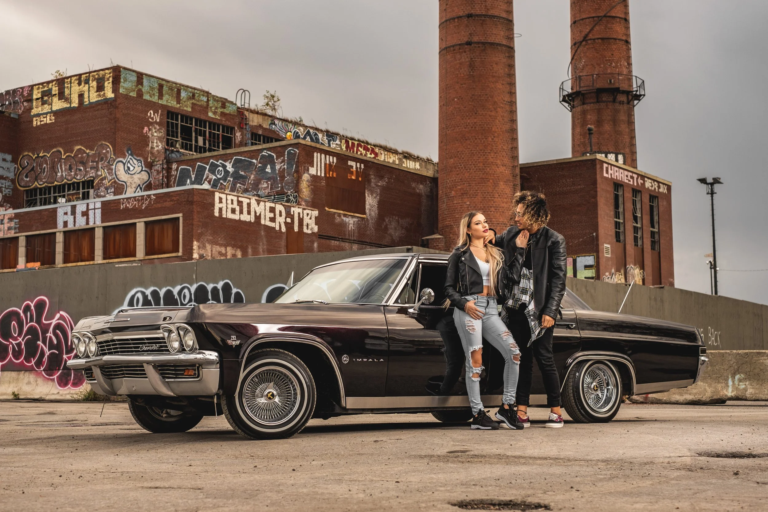 Two young women and a young man standing next to a vintage black Cadillac in front of a graffitied industrial building.