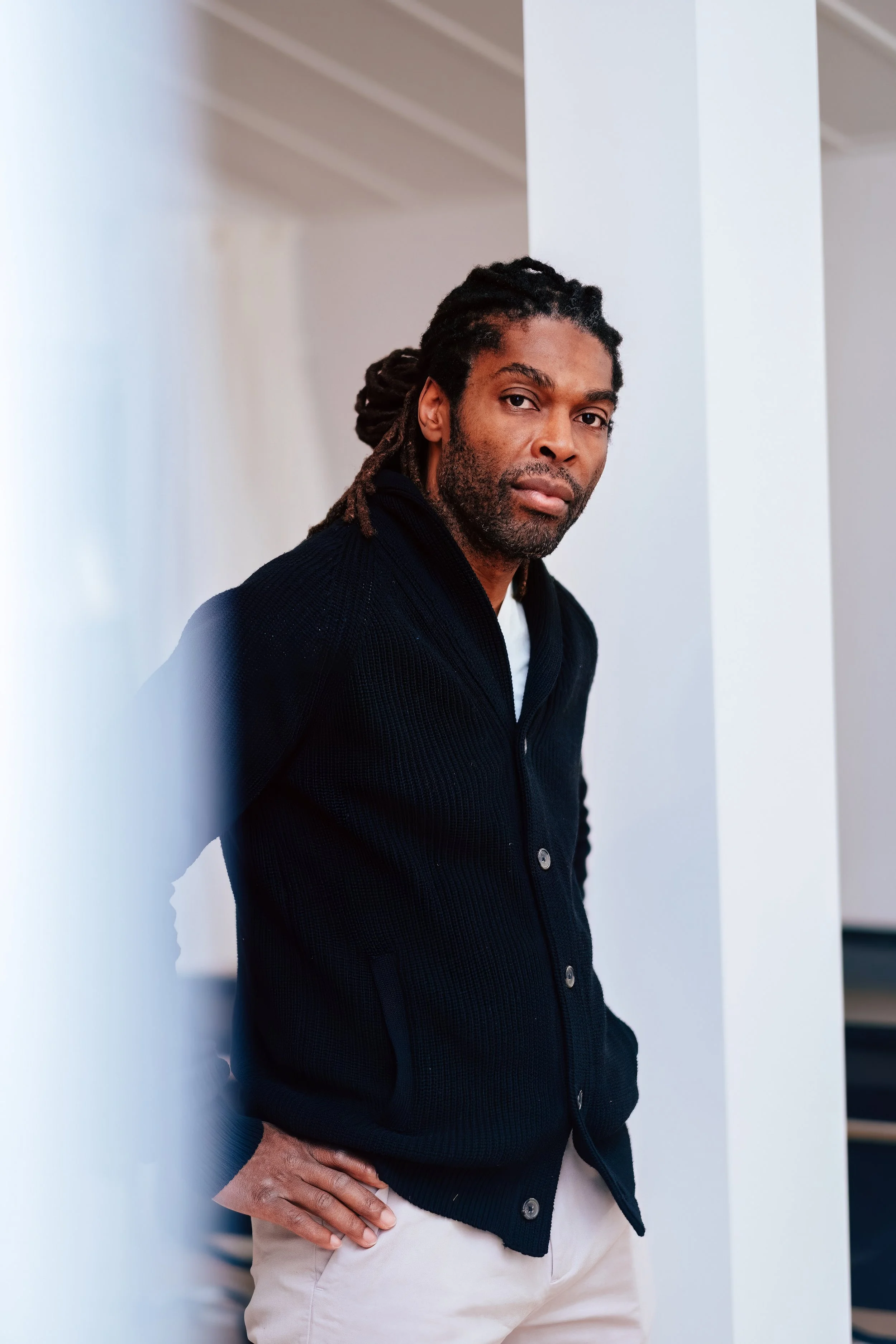 Videographer in Montreal capturing professional music videos, corporate videos, and live events. HigA man with long dreadlocks, a beard, and medium skin tone leaning against a white wall, wearing a black jacket and beige pants, looking at the camera.