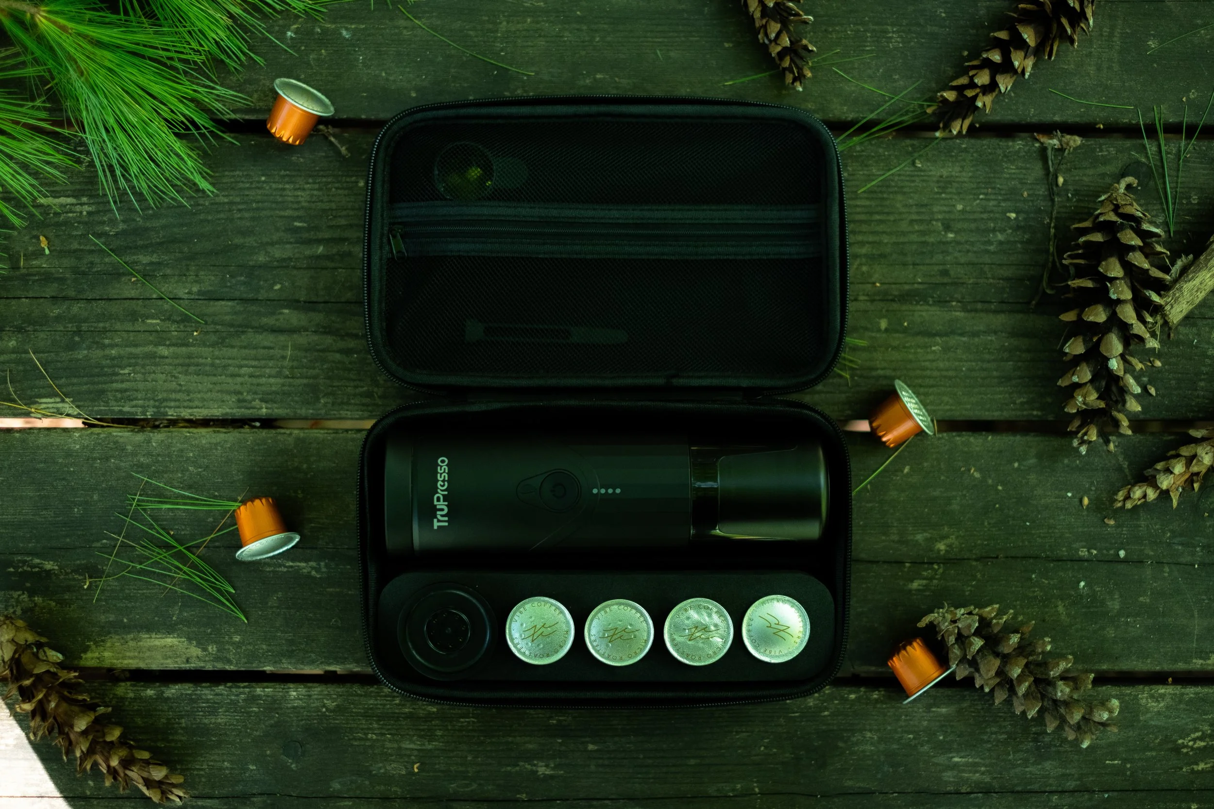 An open black case on a green wooden surface containing a flashlight and four coins, surrounded by pinecones and pine needles.