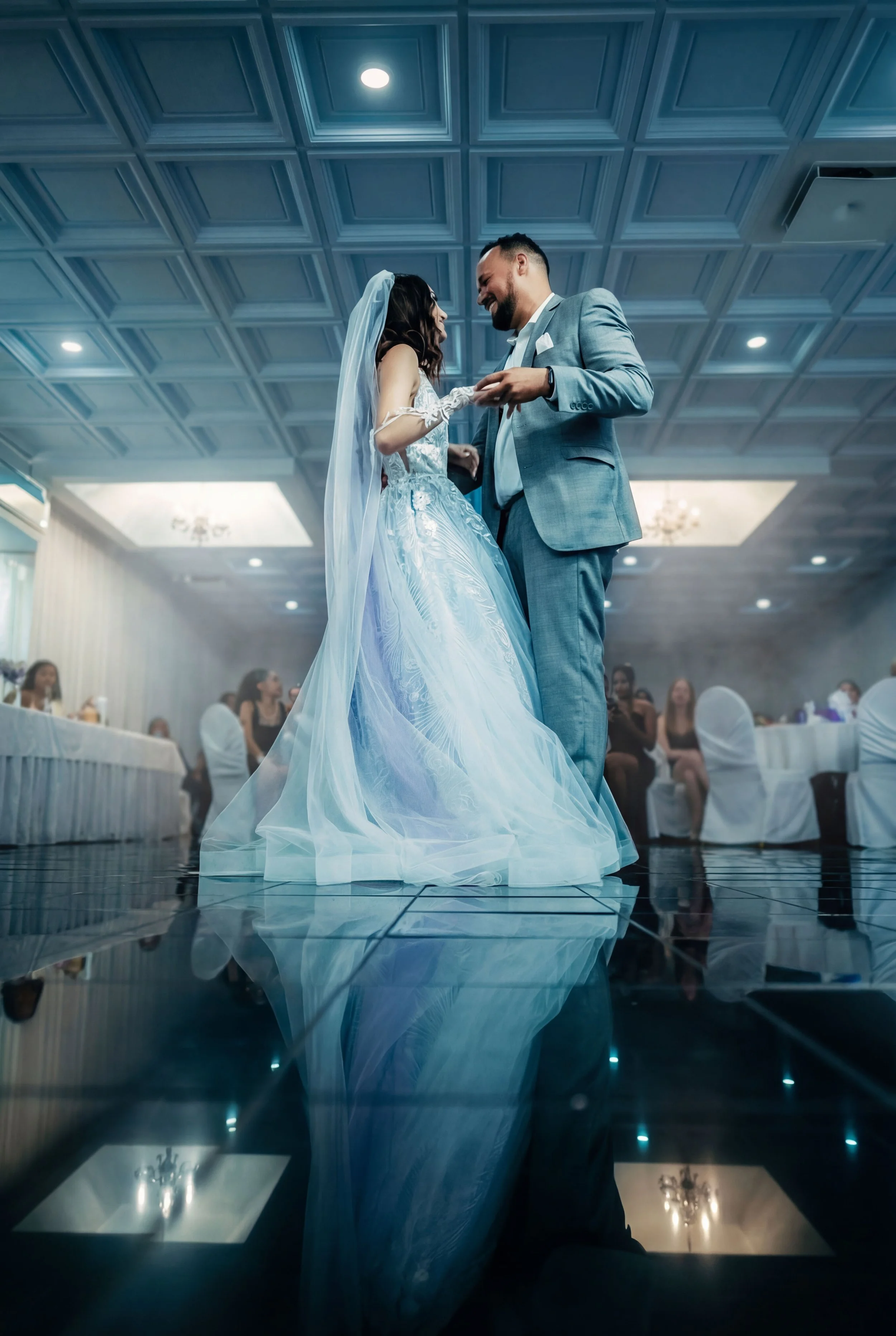 A bride and groom share a dance at their wedding reception, with guests seated at tables in the background.