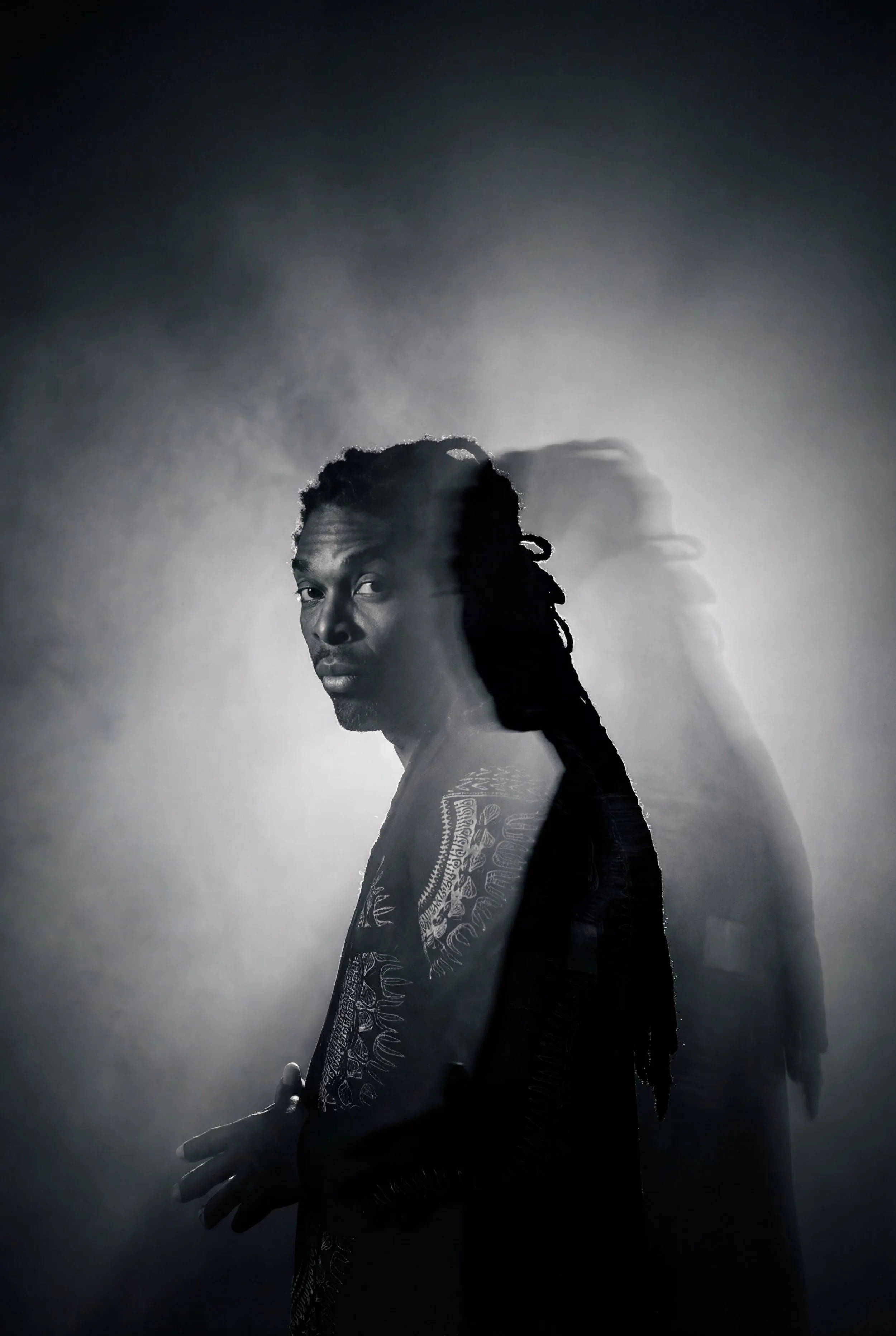 Videographer in Montreal capturing professionalA black and white photo of a man with dreadlocks, partially shirtless with tattoos on his arm, looking over his shoulder with a serious expression. The background features a shadowy duplicate of the man.