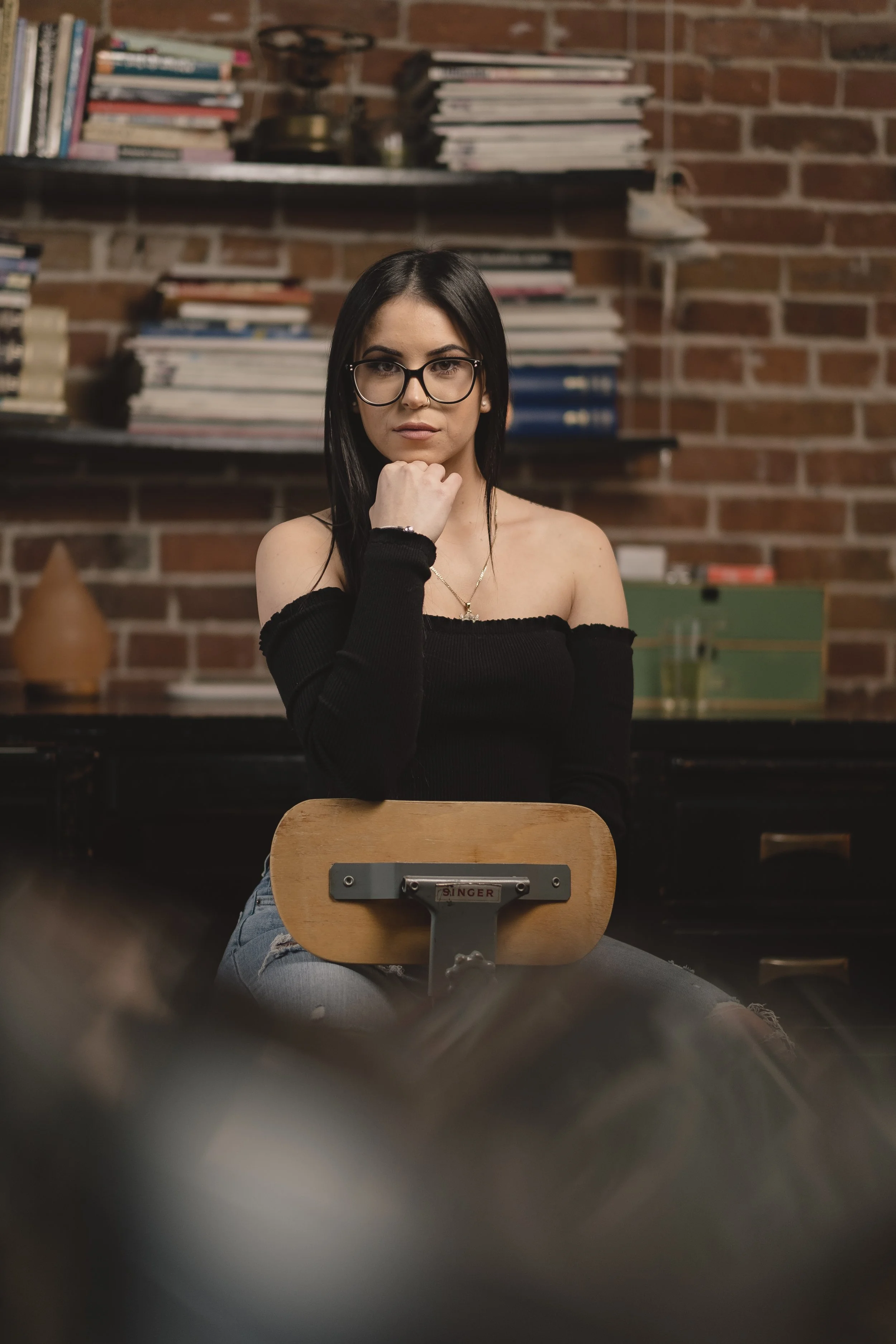A young woman with black hair, wearing glasses and a black off-the-shoulder top, sitting on a wooden chair in front of a brick wall with shelves of books.