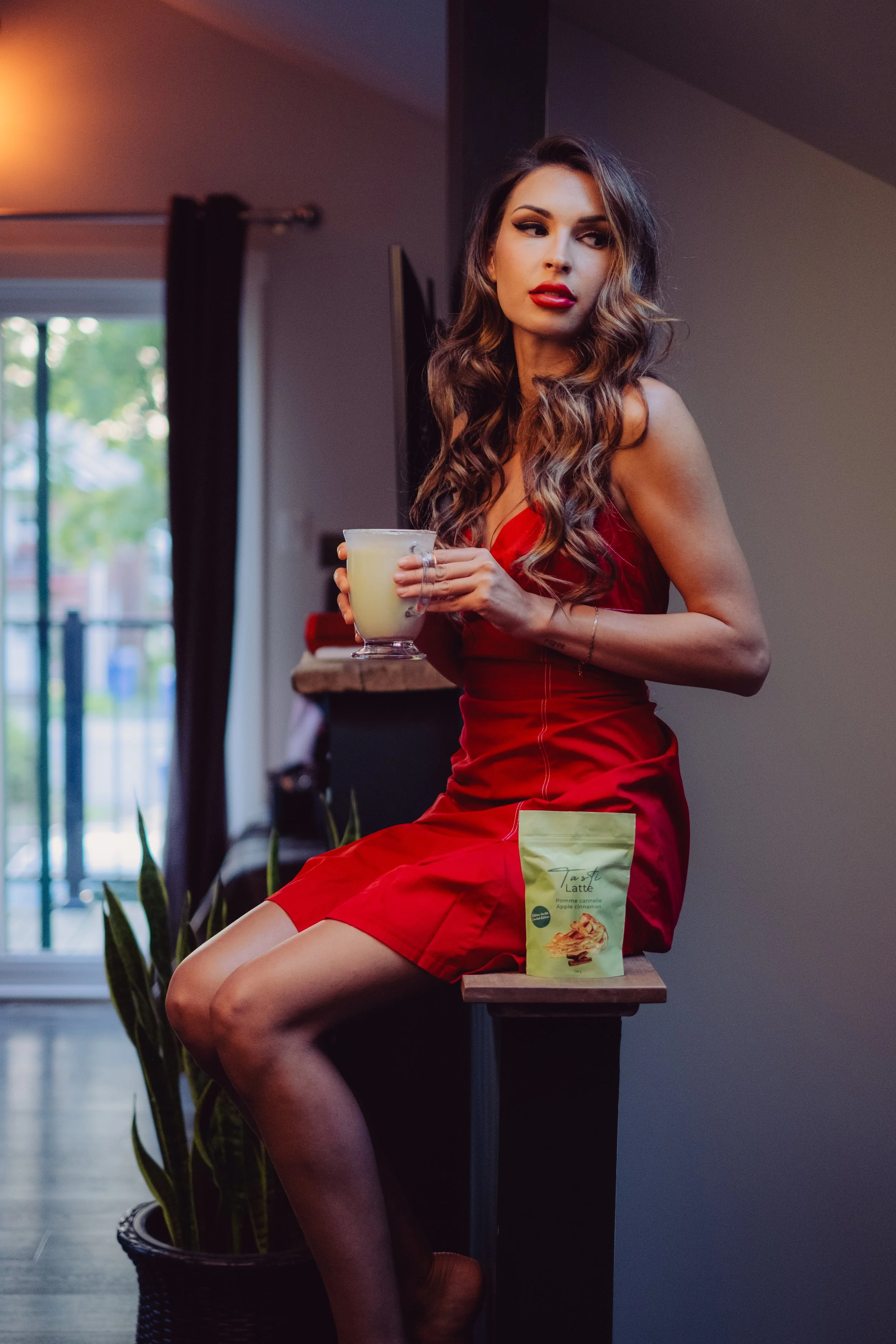 Videographer in Montreal capturing professional music videos, corpA woman with long wavy brown hair wearing a red dress sitting on a wooden counter in a living room, holding a glass of yellow beverage, with a package of cappuccino mix on the counter.