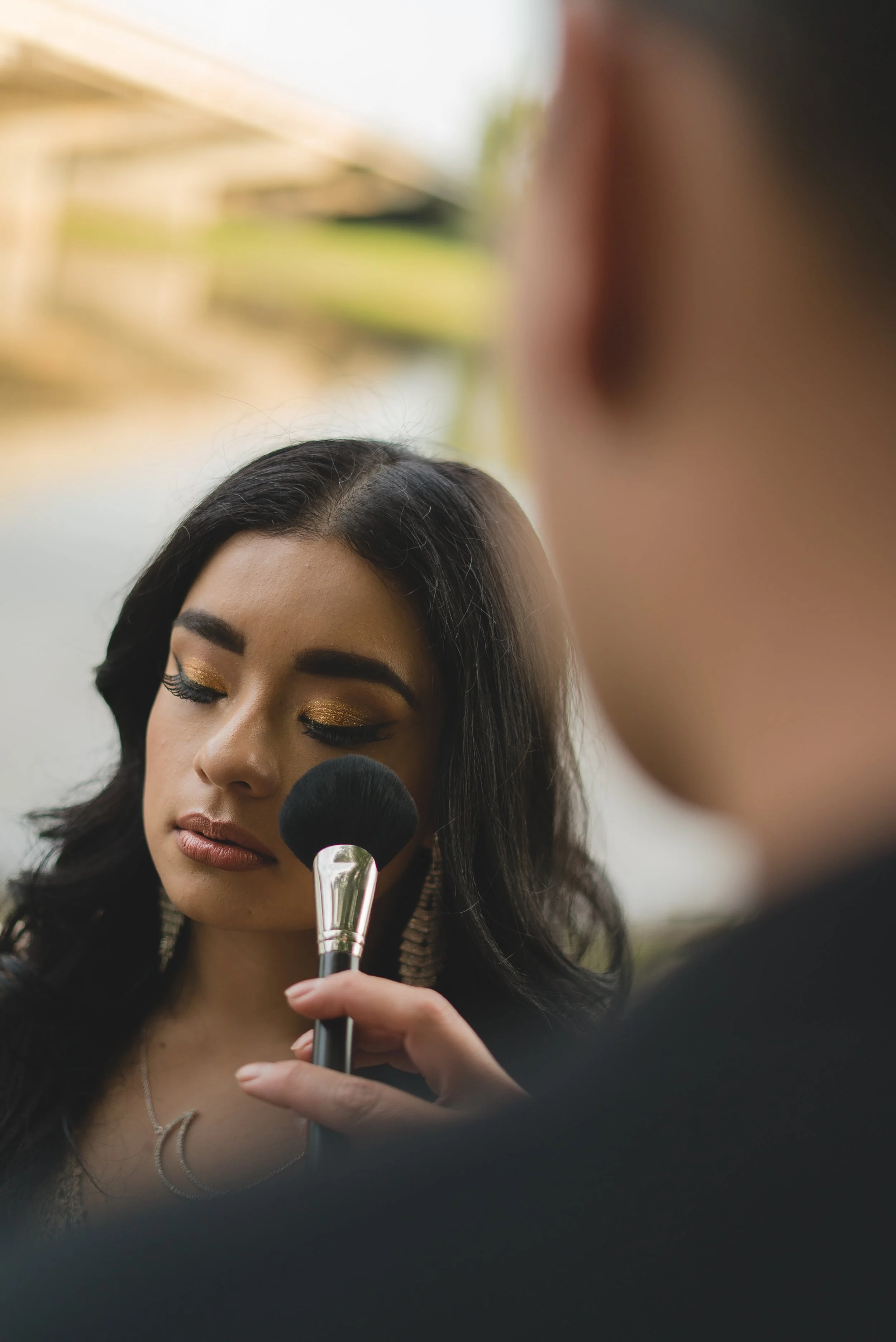 A woman with dark hair and golden eye makeup is having her face gently touched with a makeup brush held by another person.