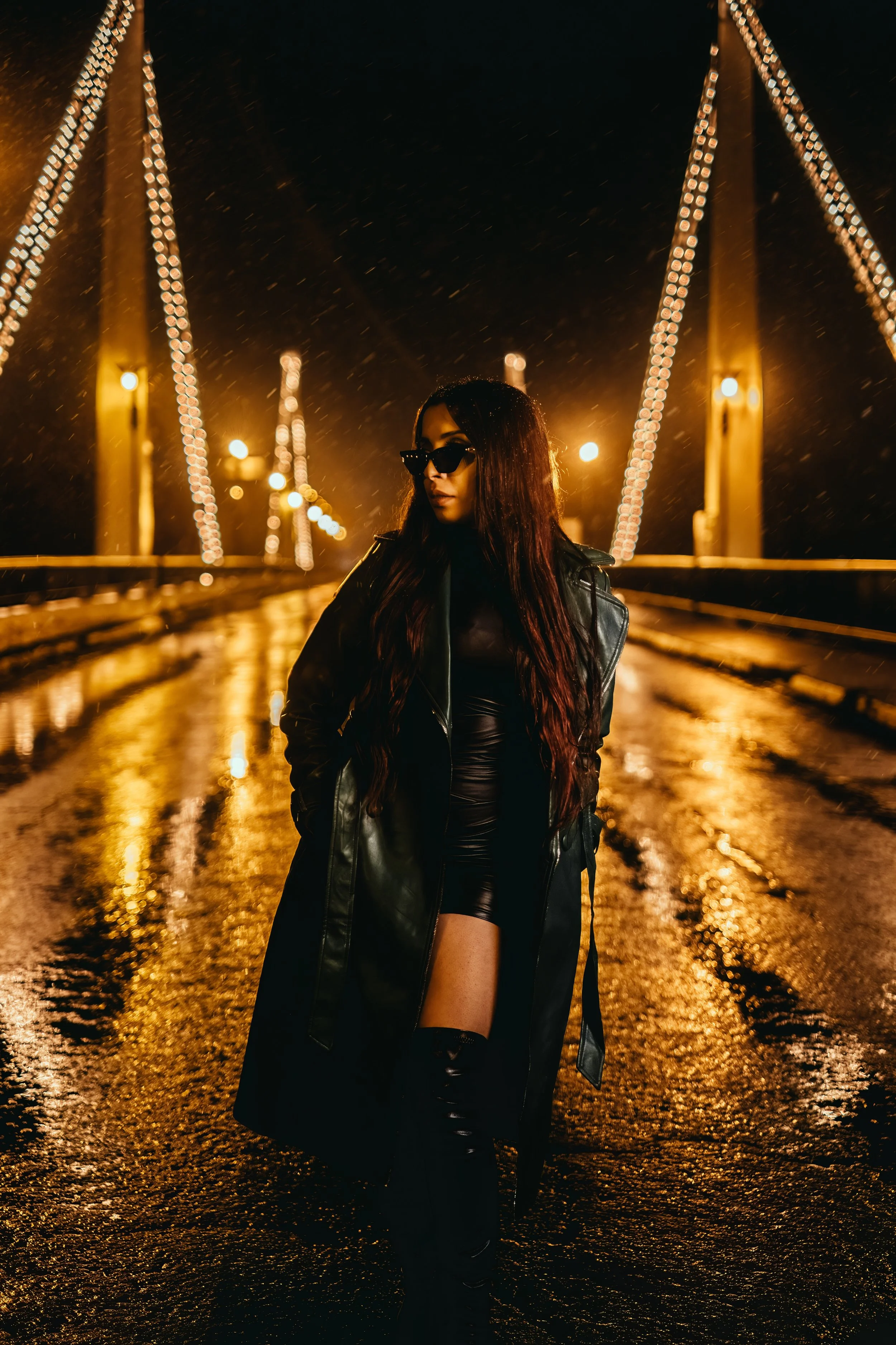 A woman in a black leather jacket, black dress, and over-the-knee boots standing on a rainy illuminated bridge at night, with streetlights and rain reflections in the background.
