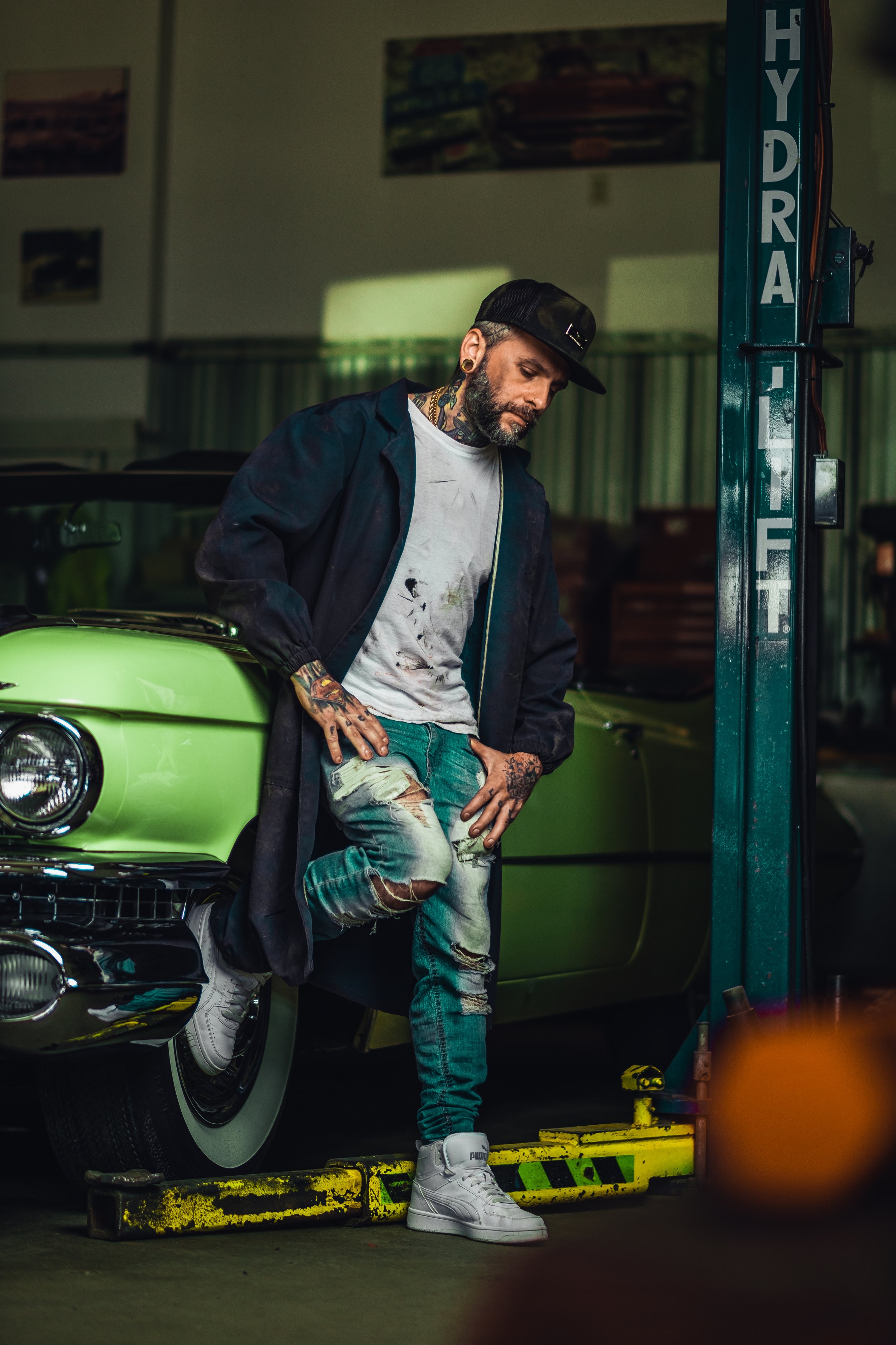 Videographer in Montreal capturing professional music videos, corporate videos, and live events. High-quality video producA tattooed man with a beard, wearing a black cap, gray t-shirt, and torn jeans, leaning against a vintage green car in a garage.