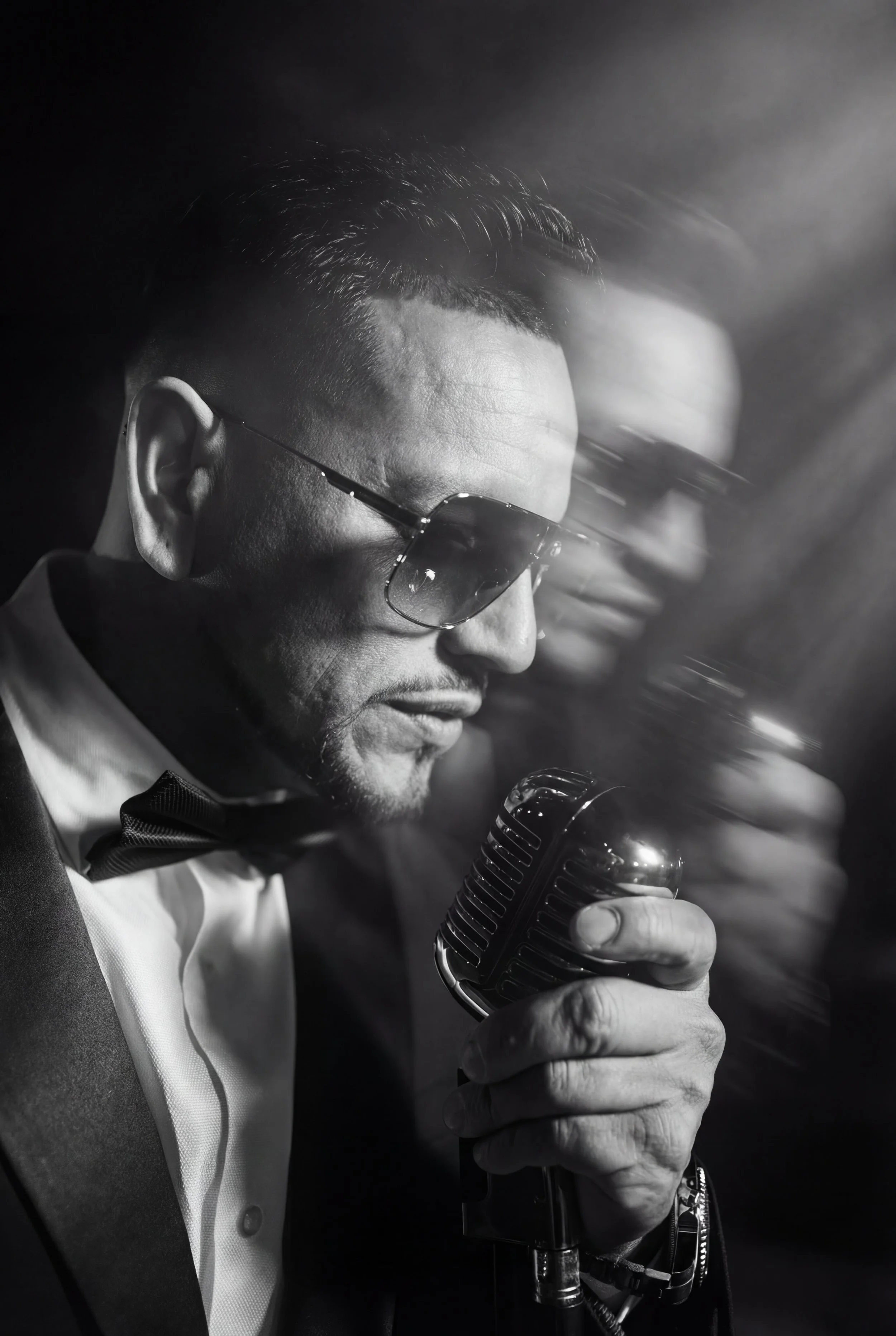 Black and white photograph of a male singer wearing sunglasses, a tuxedo, and a bow tie, singing into a vintage microphone, with a blurred second person in the background.