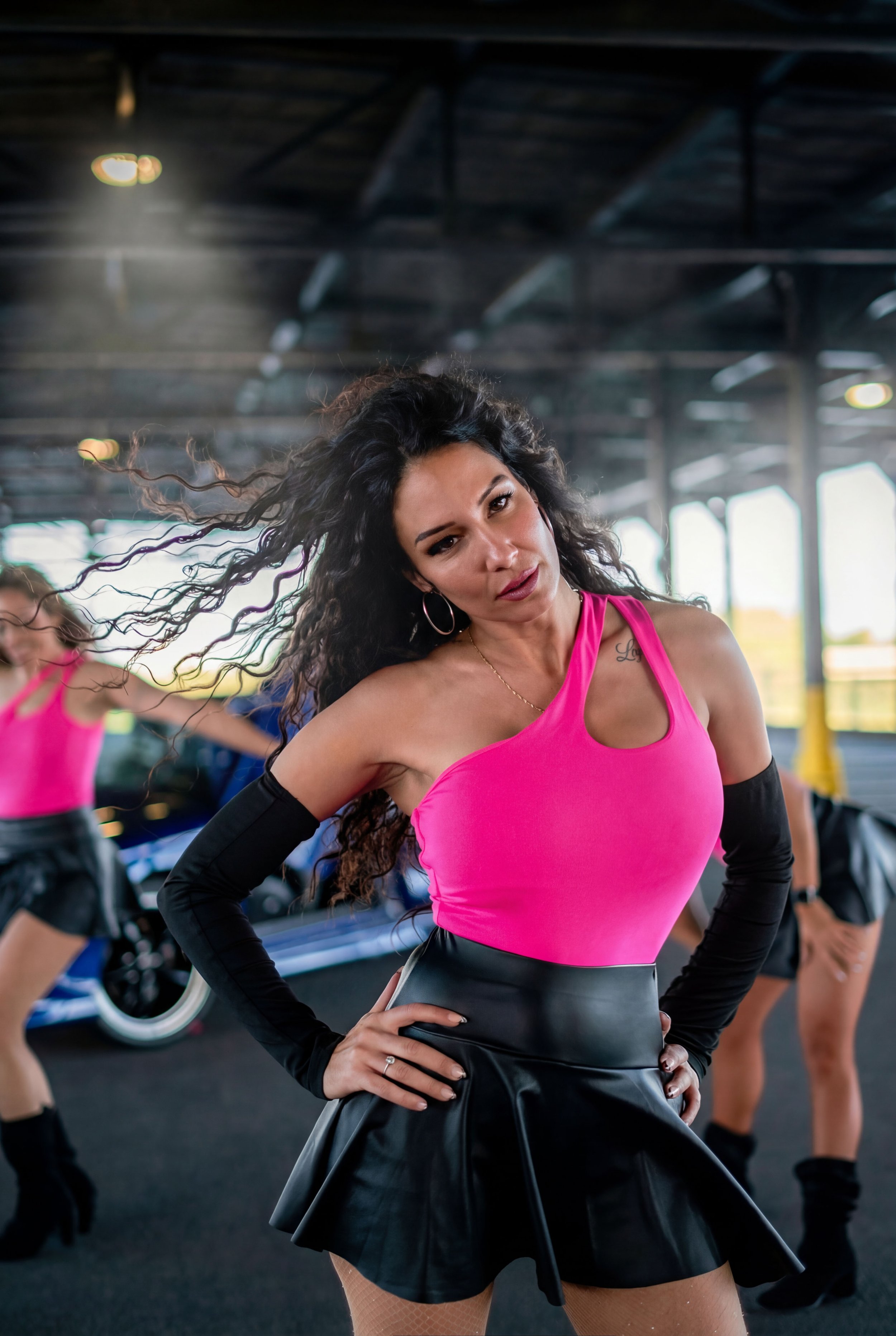 A woman with long, curly dark hair, wearing a pink sleeveless top, a black leather skirt, and black arm sleeves, is posing with one hand on her hip inside a gym. Other women are dancing or exercising in the background.
