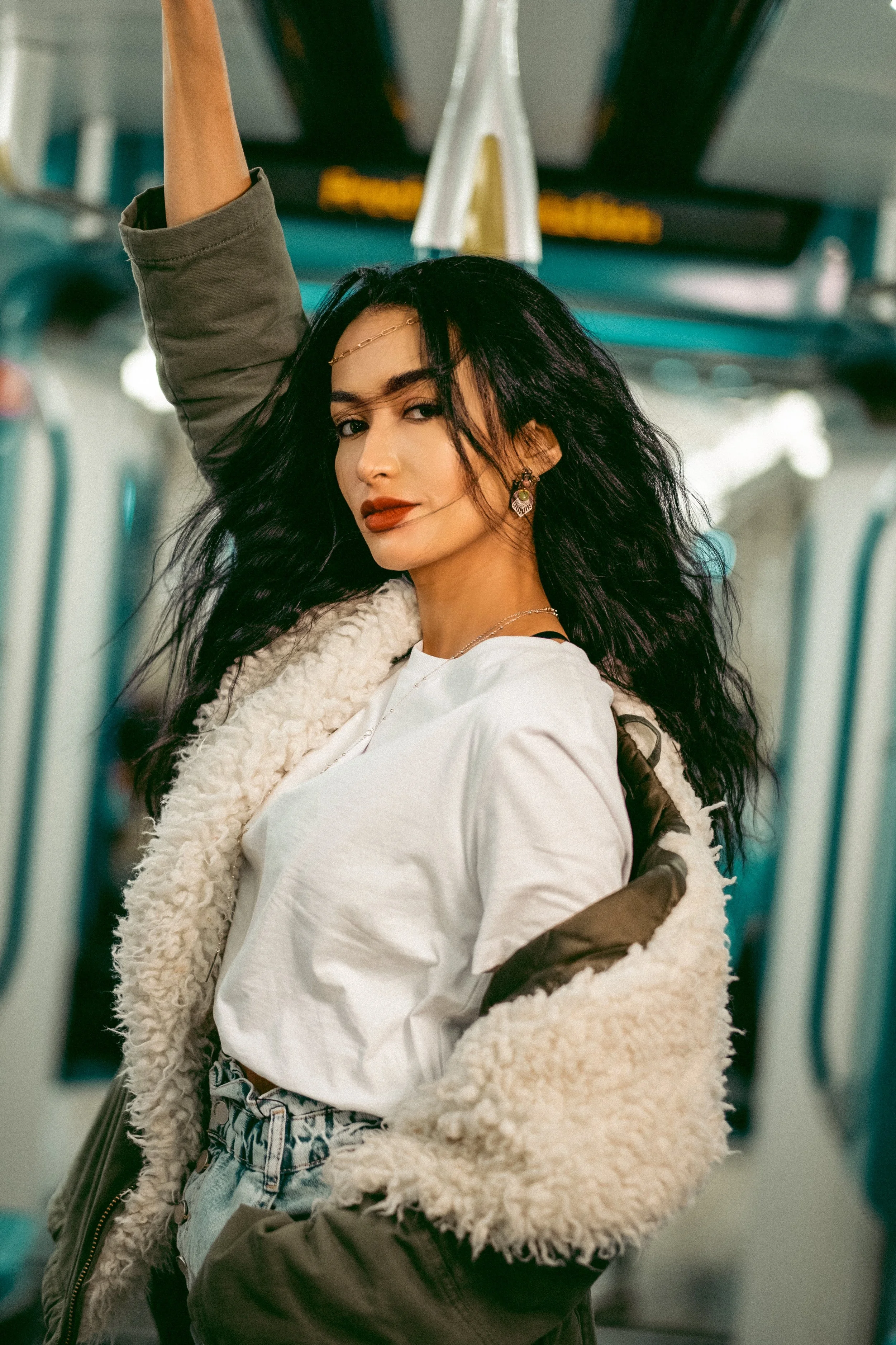 A woman with long black hair, wearing makeup and earrings, standing inside a public transportation vehicle, possibly a bus or train, with her arm raised holding a handrail.