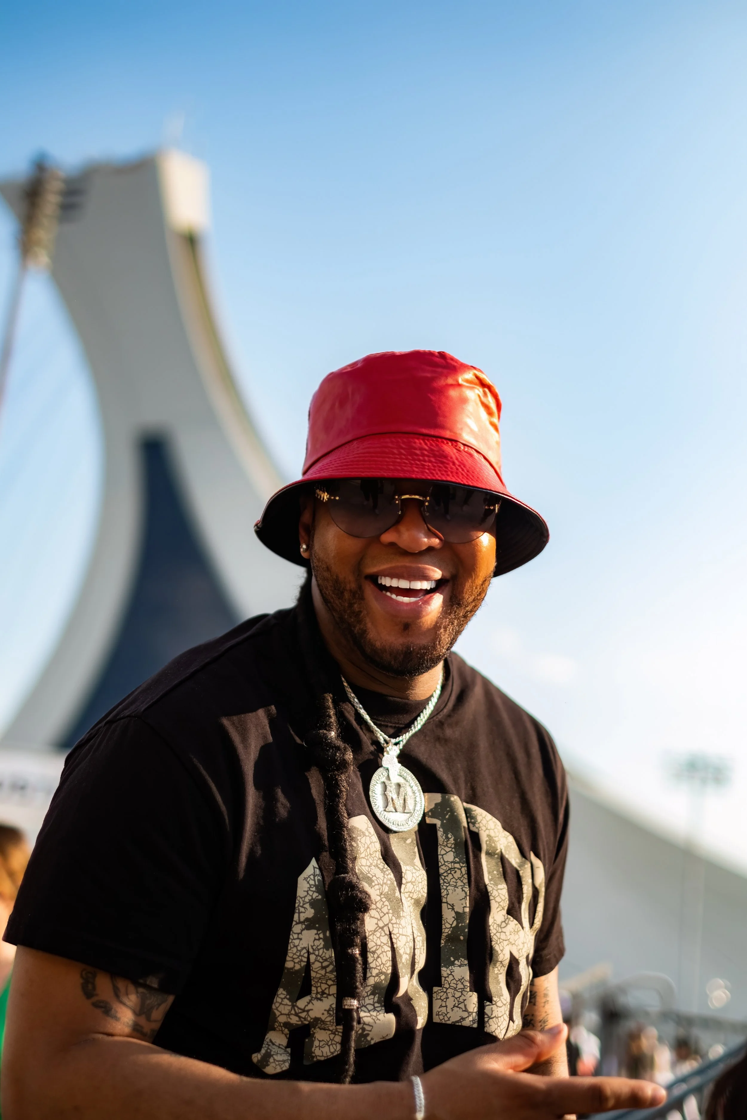 A smiling man wearing a red bucket hat, sunglasses, and a black T-shirt with gold-colored text, standing outdoors with a modern white architecture structure and a clear blue sky in the background.