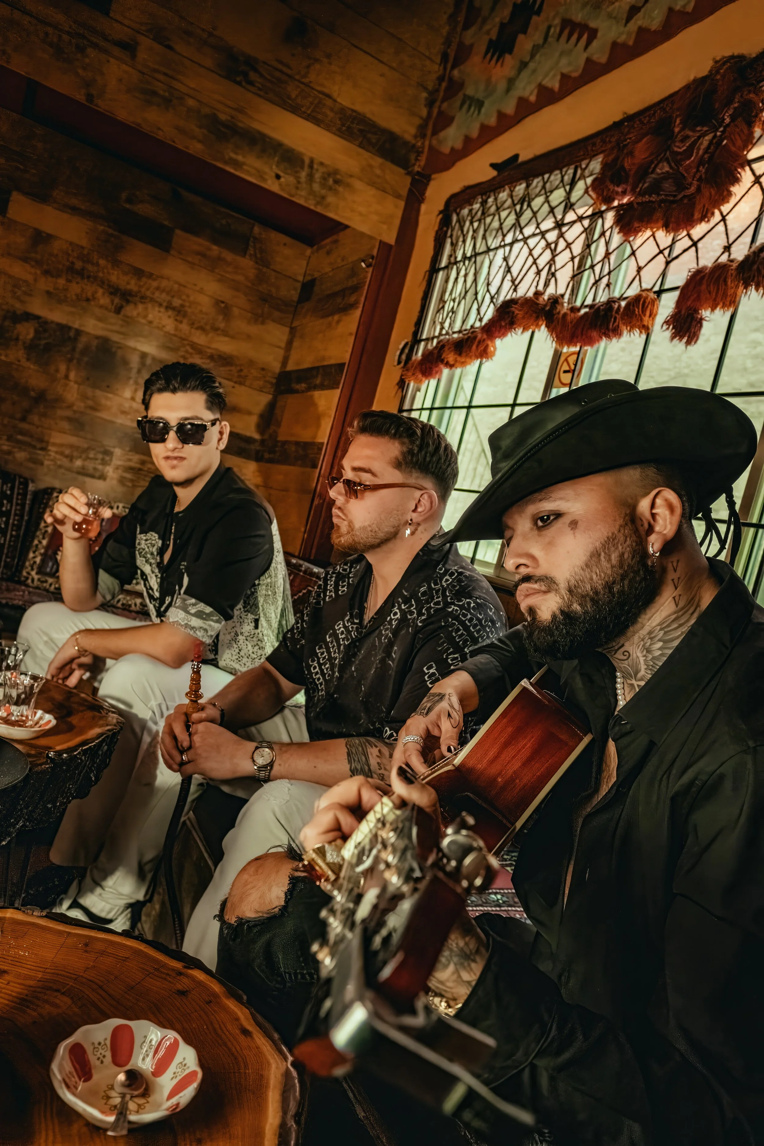 Three men sitting in a rustic room, one playing guitar, another holding a hookah, and the third holding a glass of drink. The room features wooden walls, a window with a decorative curtain, and a small ceramic bowl with a spoon in the foreground.