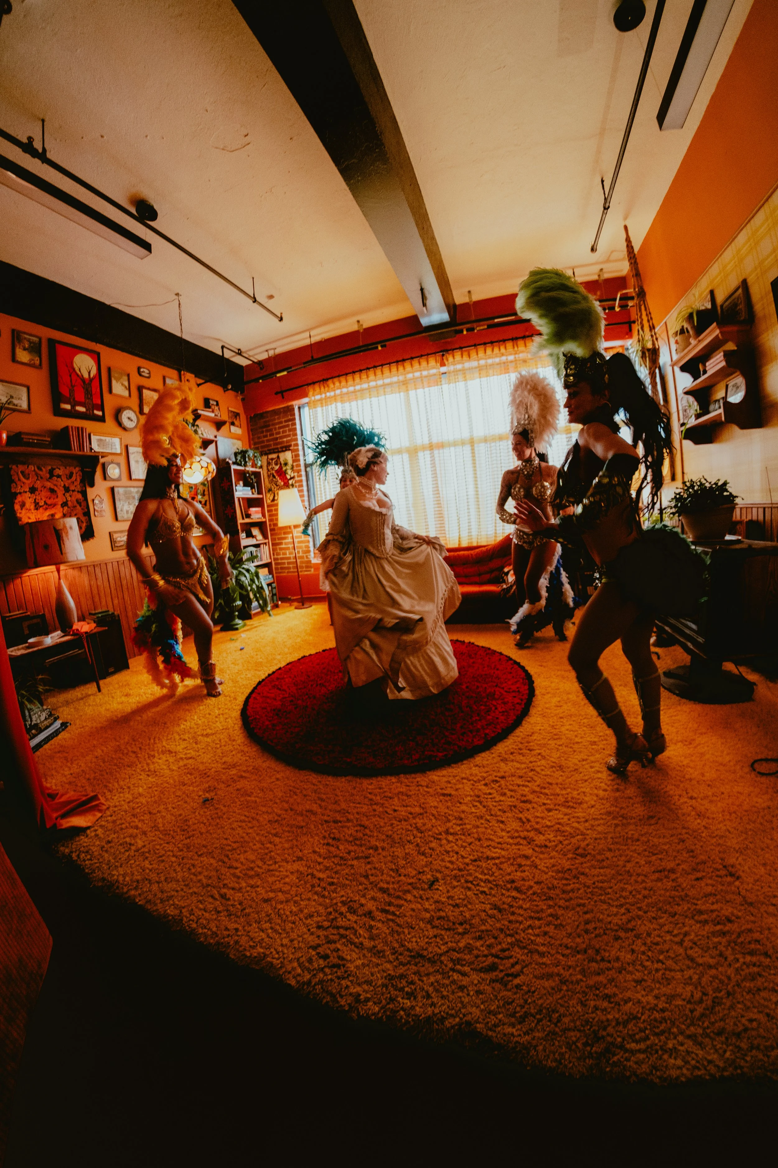 Women in colorful, feathered costumes dancing in a vintage-style living room with orange walls, a red circular rug, and a large window with curtains.