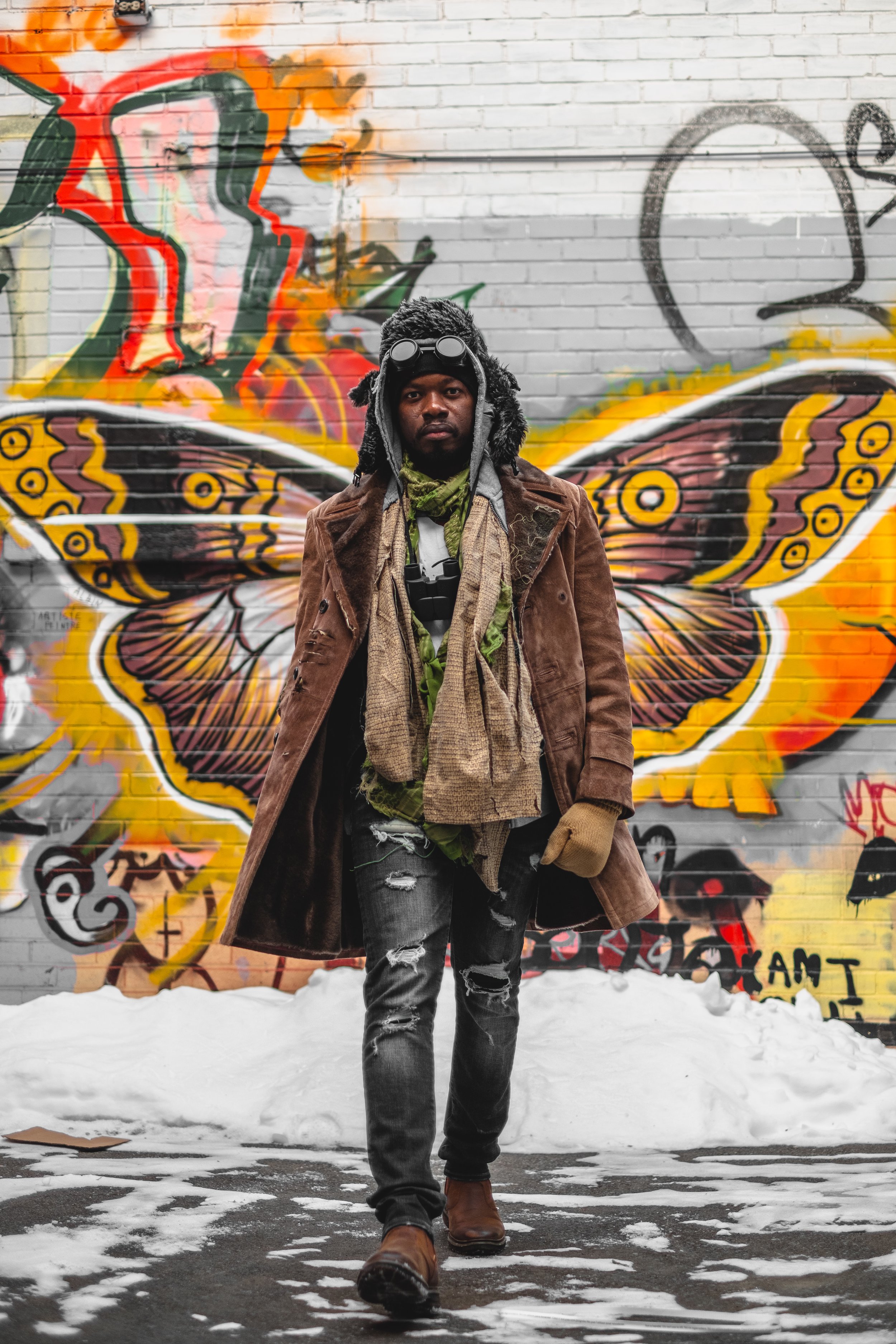 A man standing in front of a graffiti mural of butterfly wings on a brick wall, dressed in winter clothing, with snow on the ground.