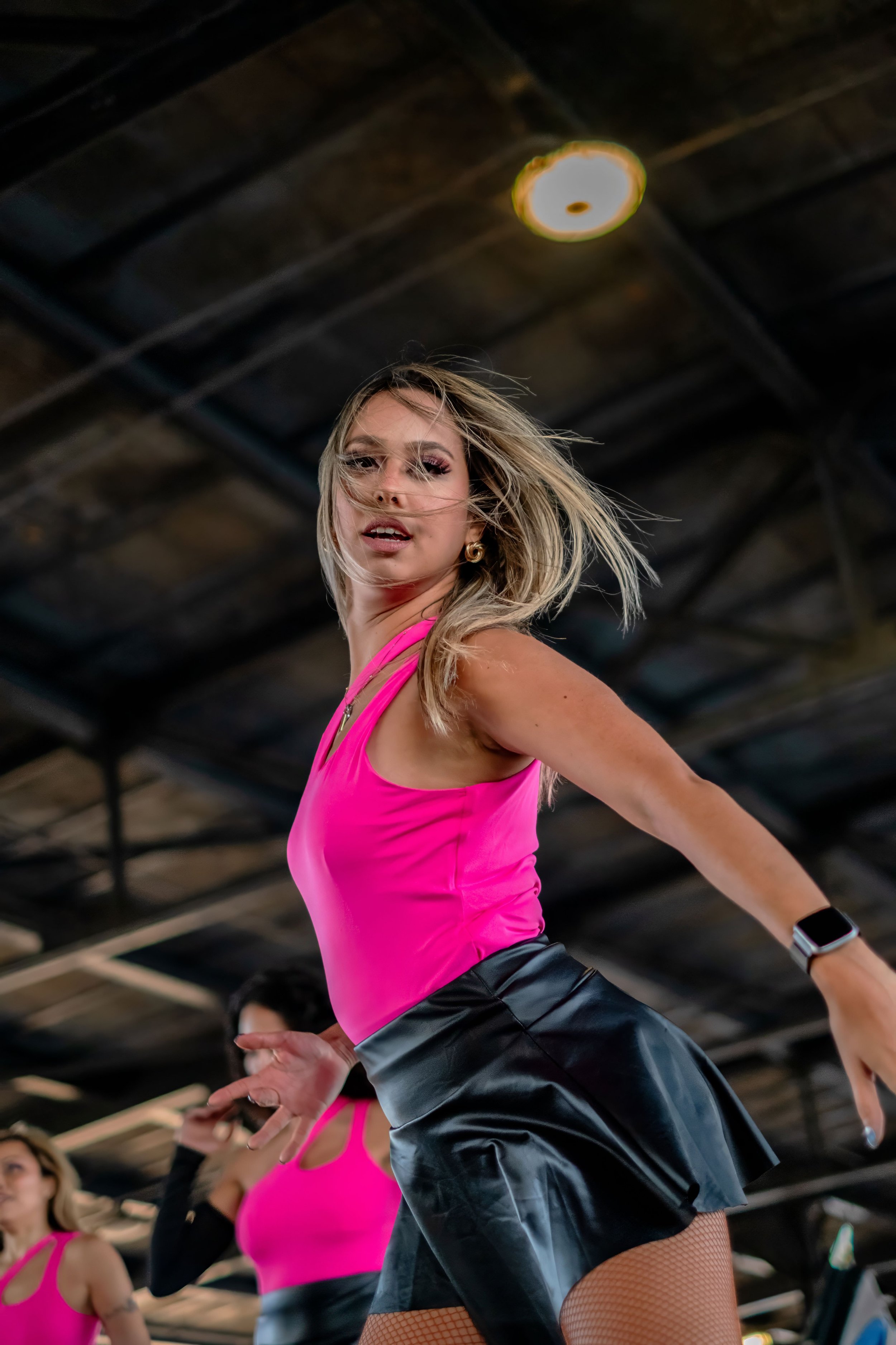 Videographer in Montreal capturing professional music videos, corporate videos, and live events. High-quality video pA woman in a pink top and black leather skirt dancing in a gym with other women in the background under a ceiling with exposed beams.