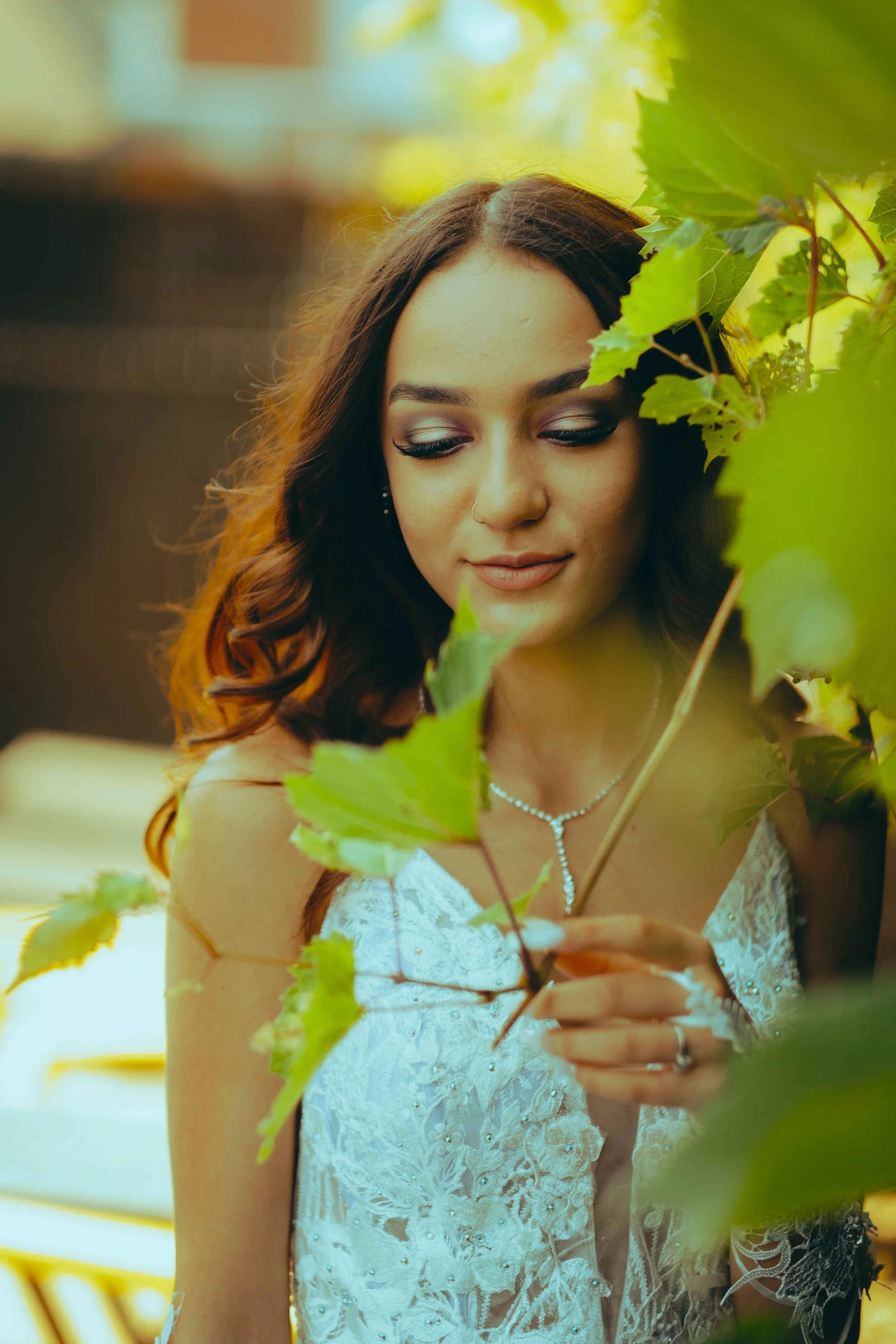 A woman in a white dress with lace details, jewelry, and makeup, standing among green leaves outdoors, gently holding a branch with leaves, with a soft background.