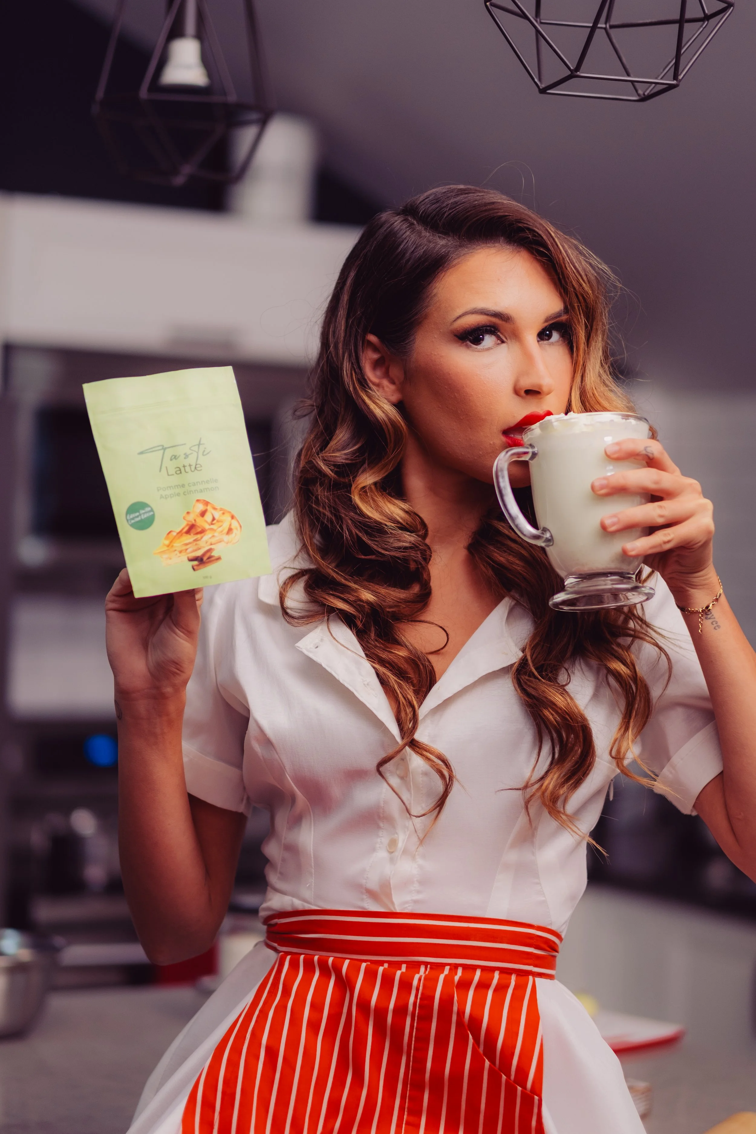 A woman with long, wavy brown hair sipping from a large glass mug of a creamy drink, holding a packet of apple cinnamon flavored latte in her other hand, in a kitchen with modern decor.