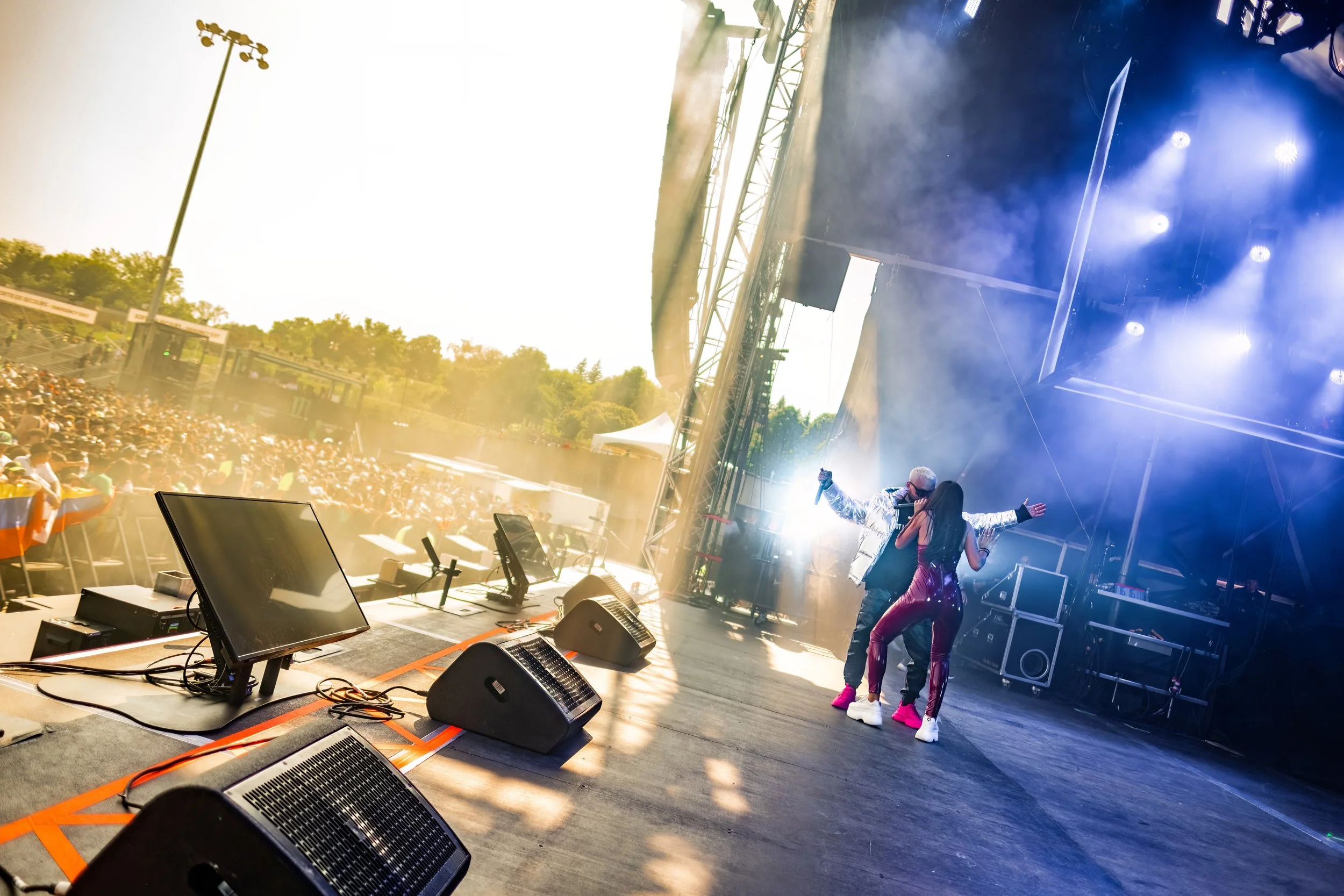 Two performers singing on stage during a concert with a large crowd in the background.