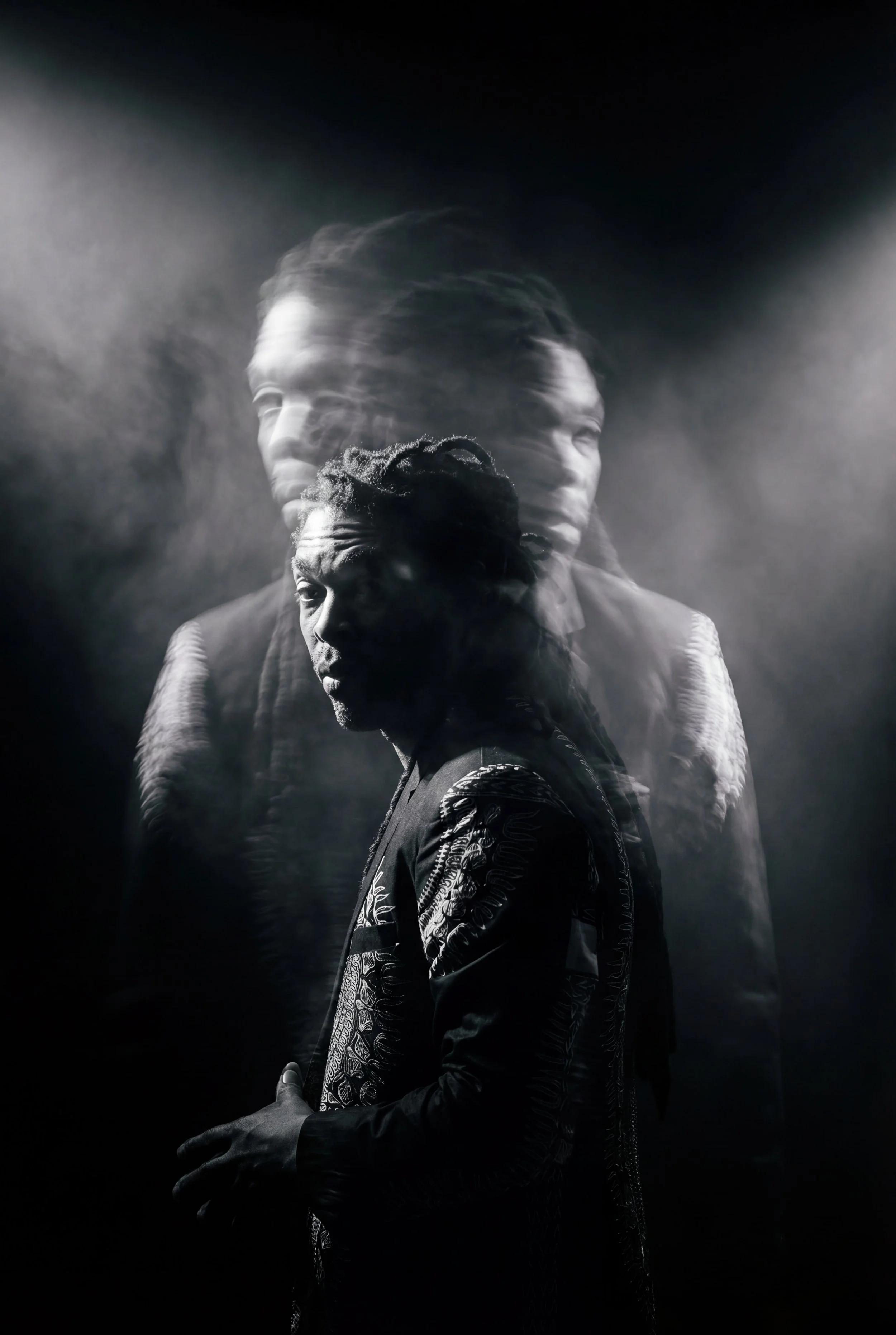 Black and white photo of a man with dreadlocks and patterned jacket, with a blurred duplicate image behind him creating a motion effect.